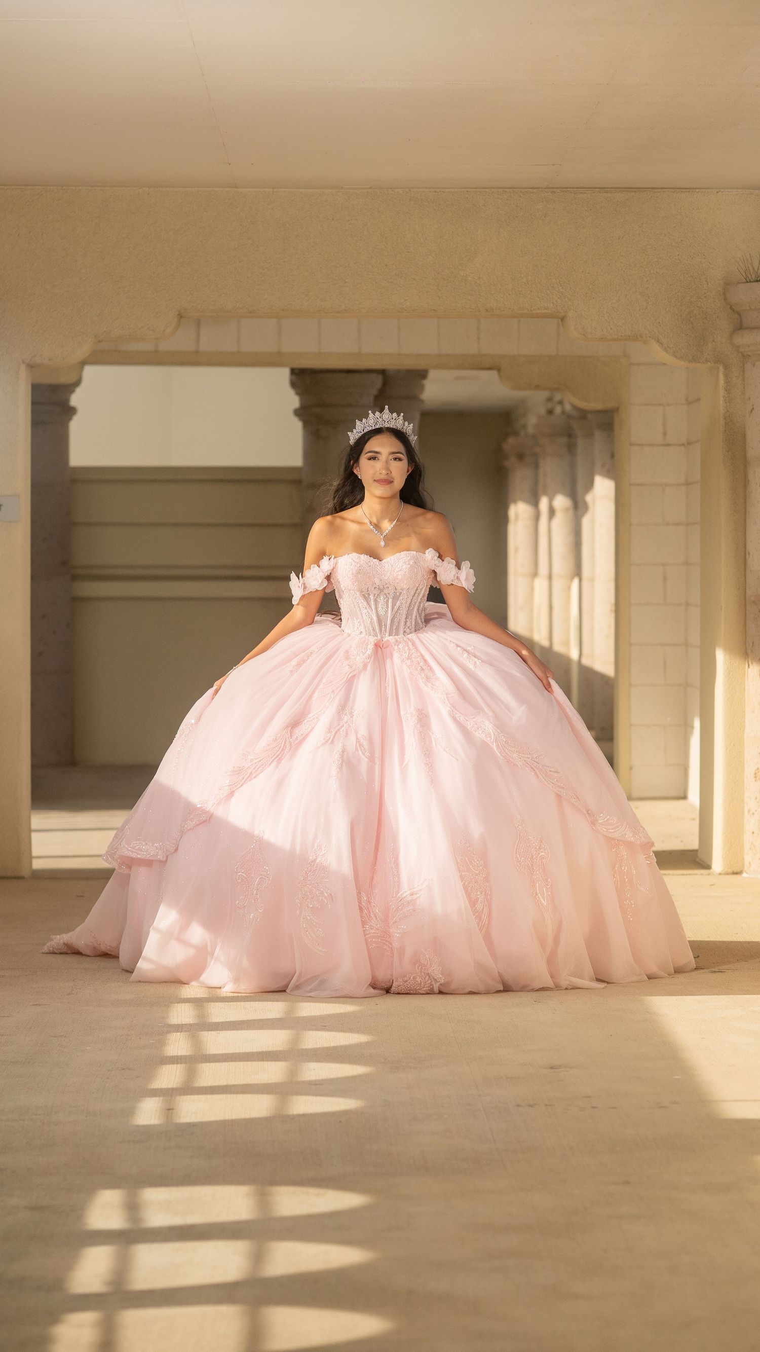 Elegant quinceañera in blush pink layered ball gown and crystal tiara stands in golden sunlit hall.