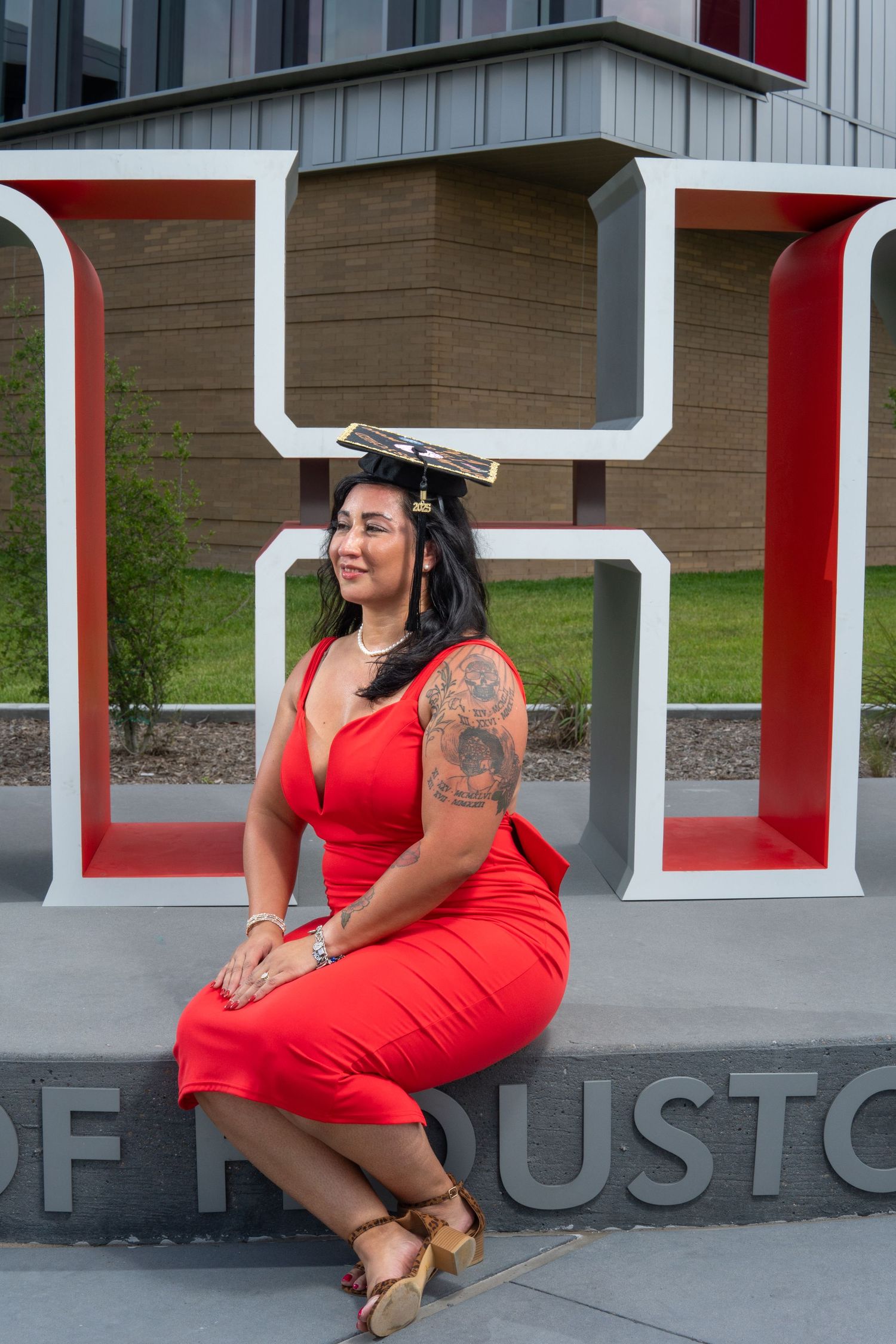 A graduate in red dress and mortarboard poses by a large university logo on a sunny day.