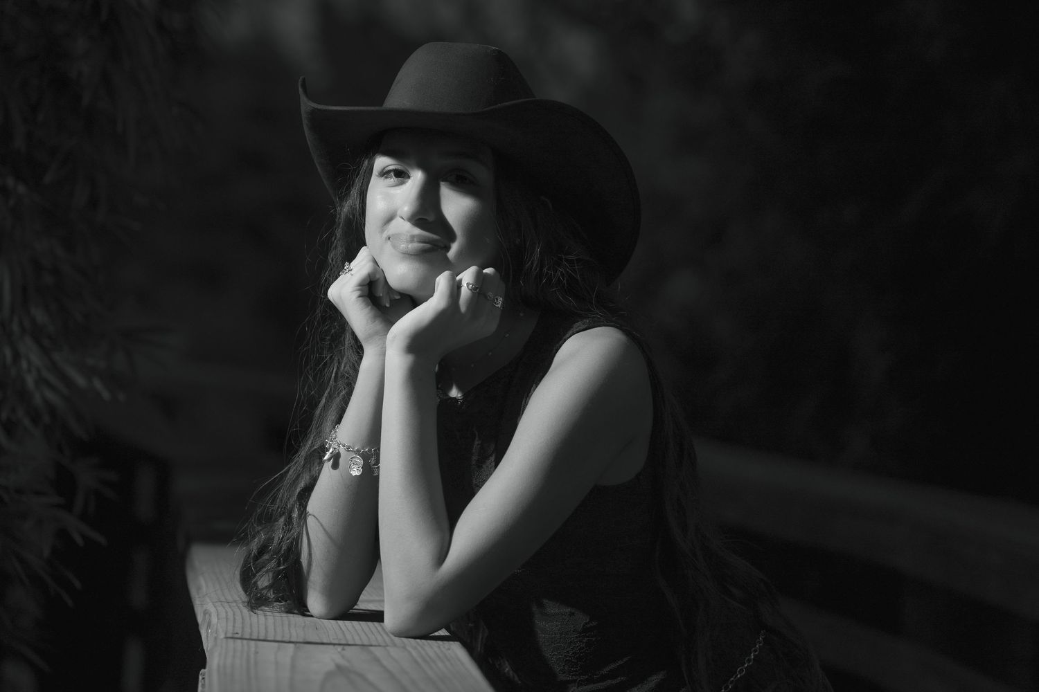 Black and white portrait of a smiling young woman in a cowboy hat, resting her chin on her hands outdoors.