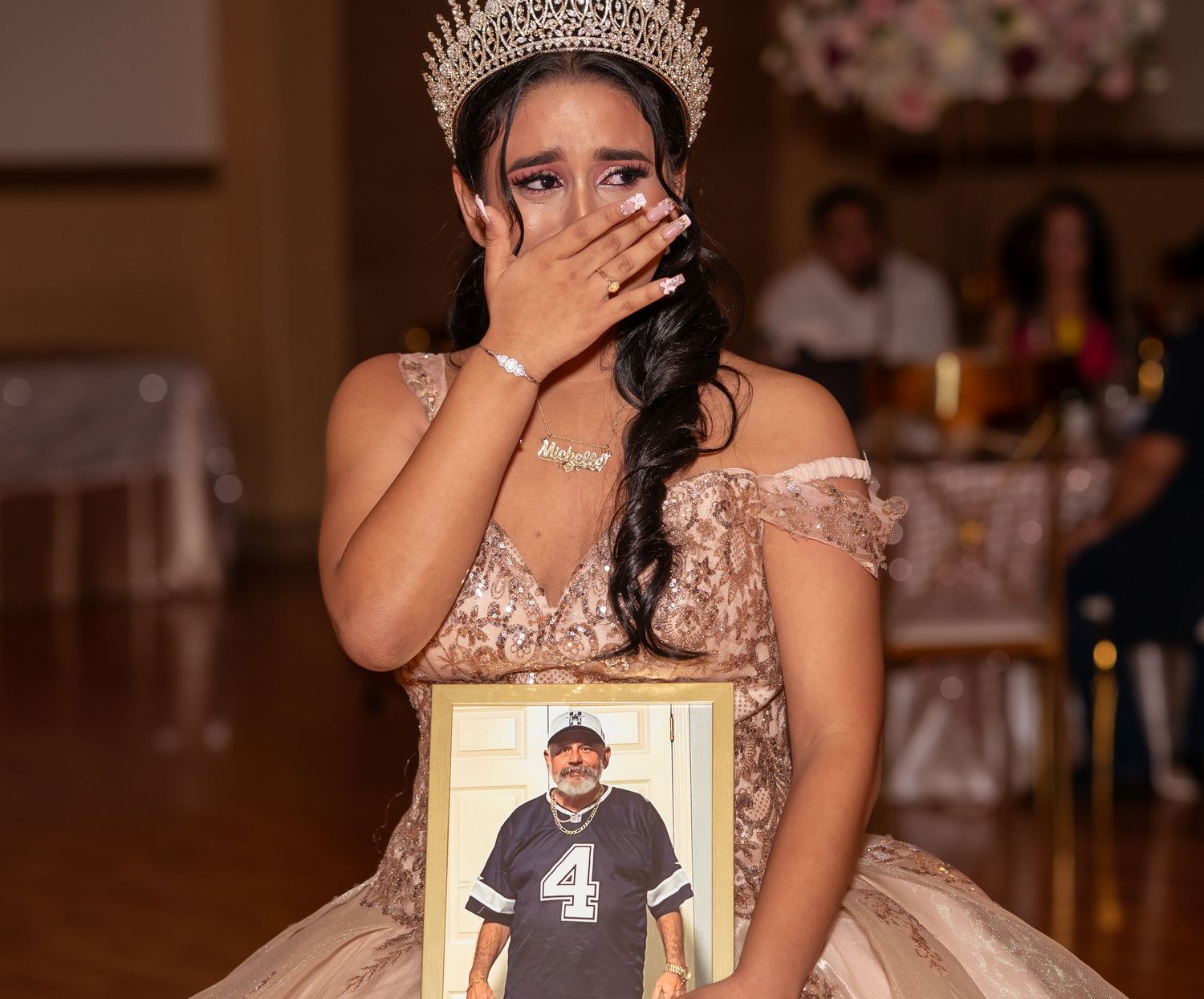 Emotional quinceañera in a gold gown and crown holds a framed photo while covering her mouth at her birthday celebration.