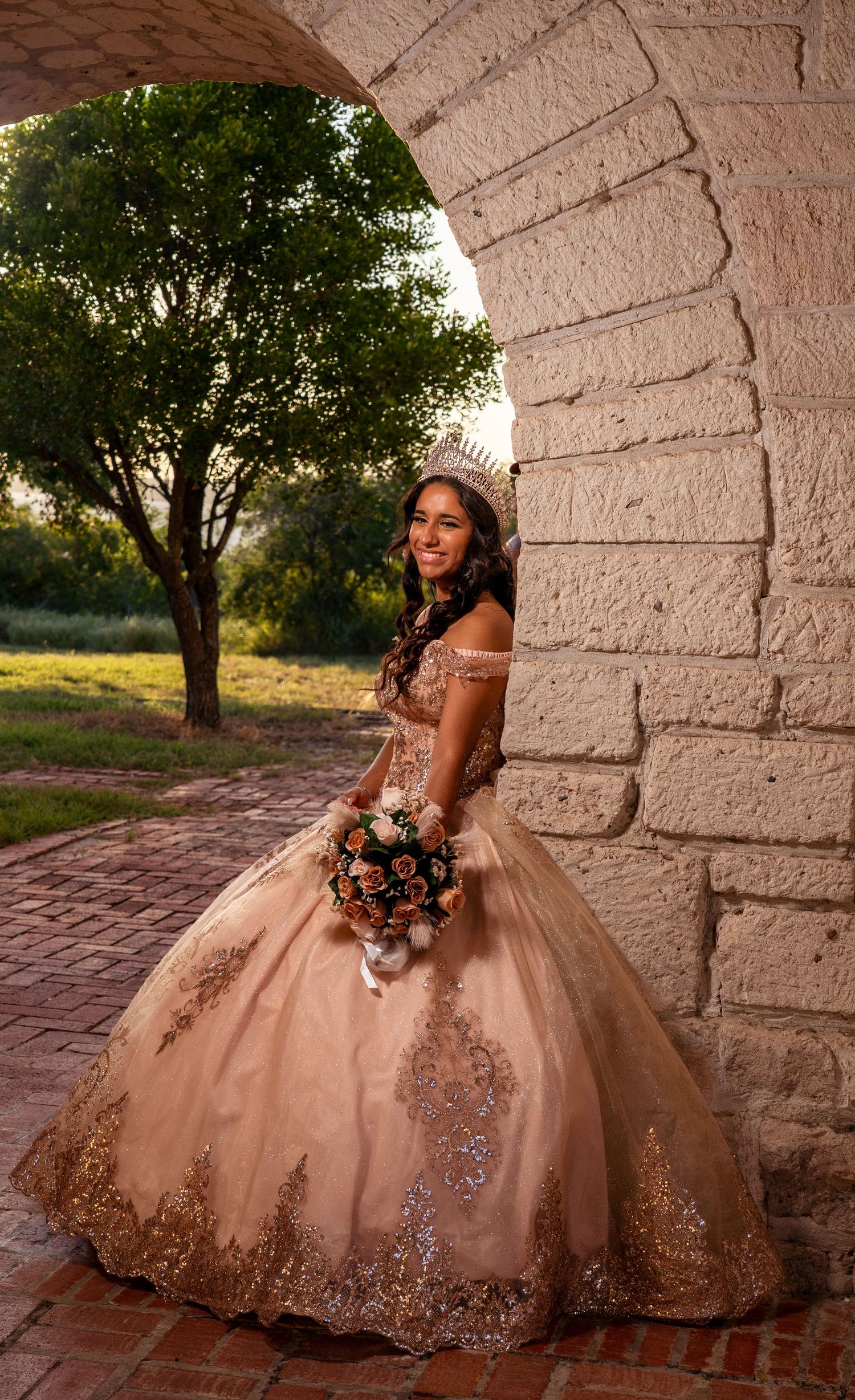 An elegant ball gown portrait featuring ornate beadwork and a tiara under a stone archway at sunset.