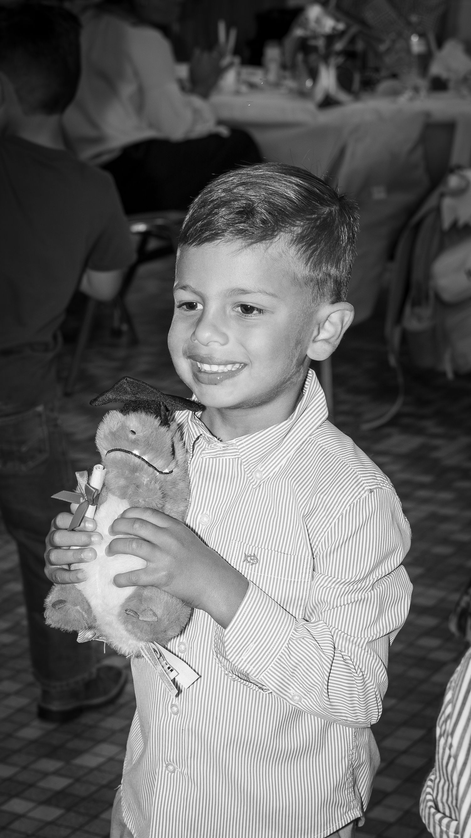 A smiling young boy in a striped shirt holds a stuffed animal toy at what appears to be a formal event.