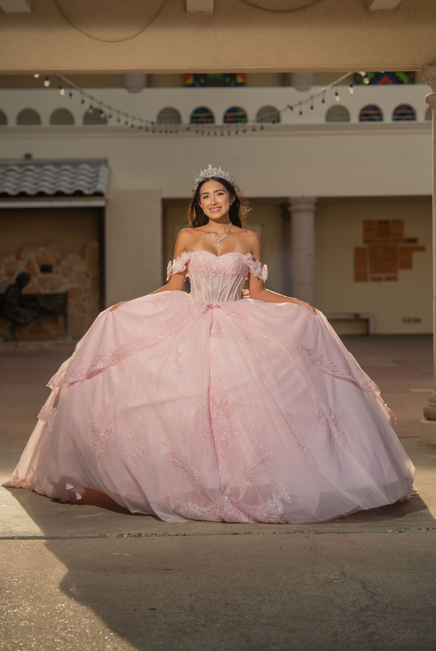 Young woman in a pink off-shoulder quinceañera ball gown with tiara poses outdoors.