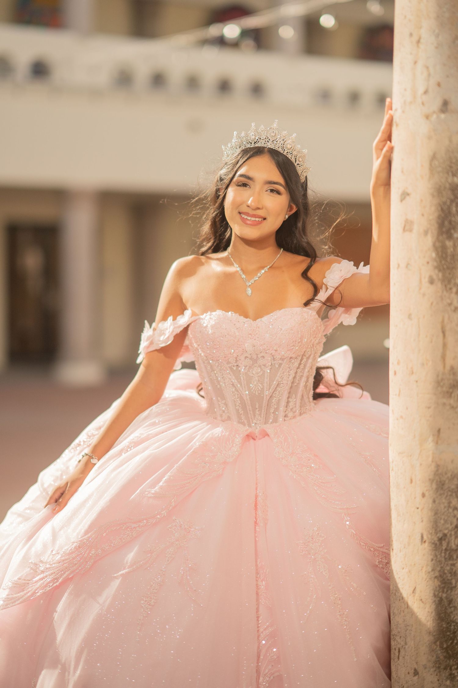 Young woman in pink quinceañera ball gown with crystal tiara poses elegantly beside a column outdoors.