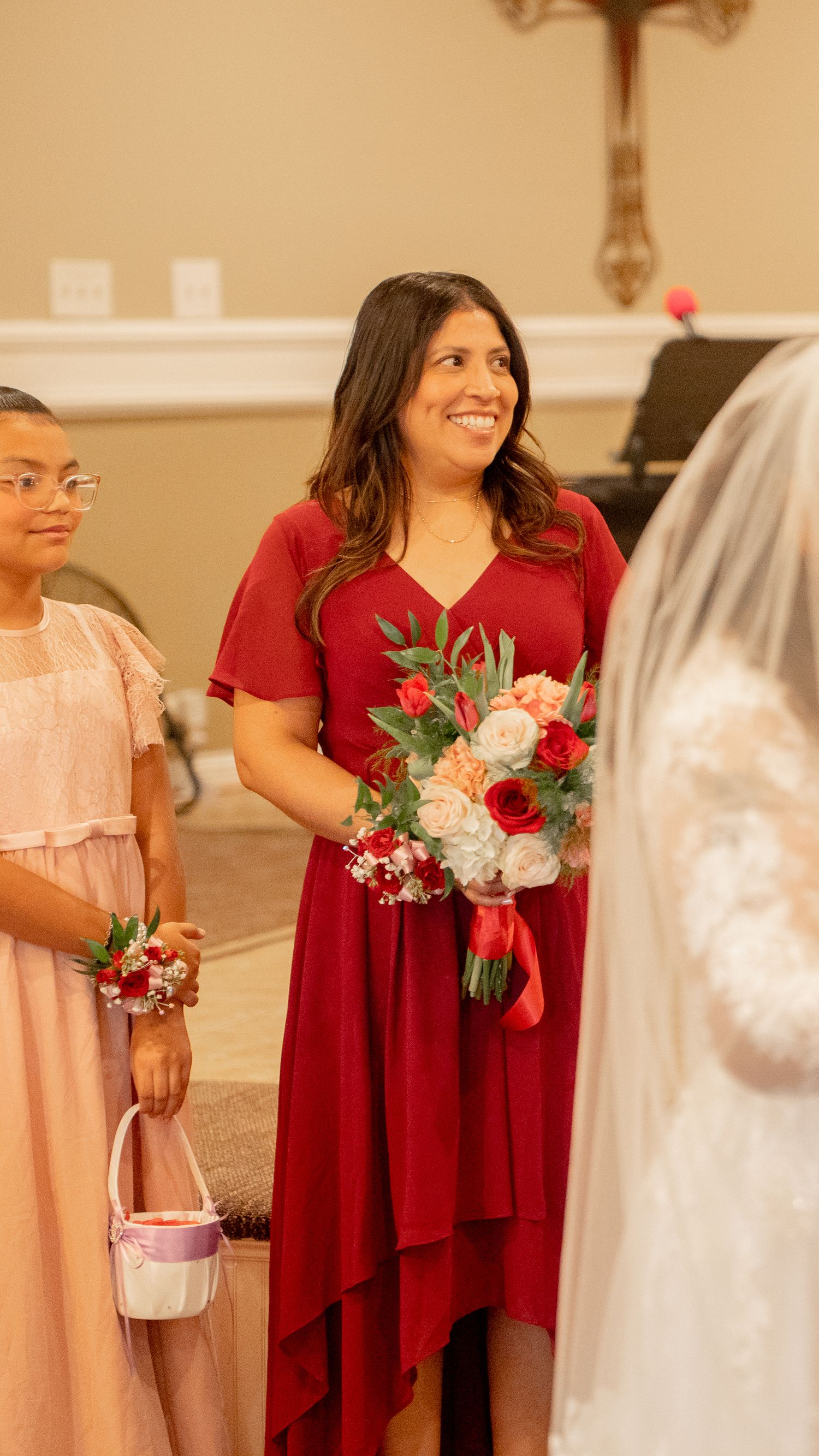 Smiling bridesmaid in red dress holding bouquet of red and pink roses during wedding ceremony.