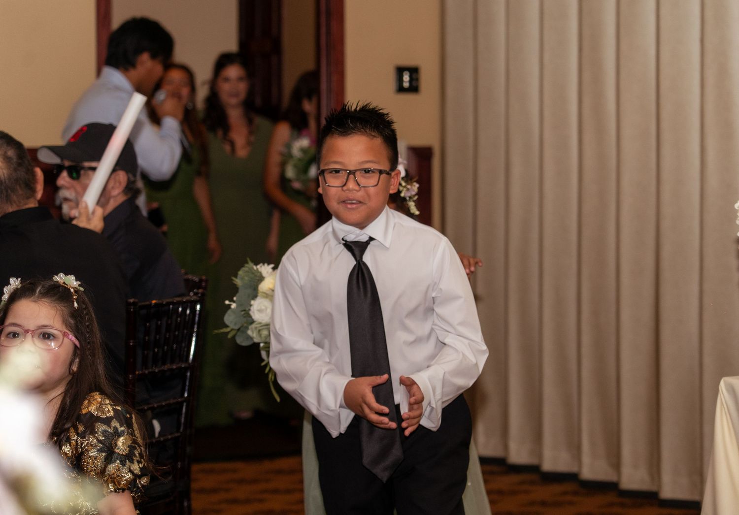 Young boy in white shirt and black tie walks down aisle at formal wedding reception event.