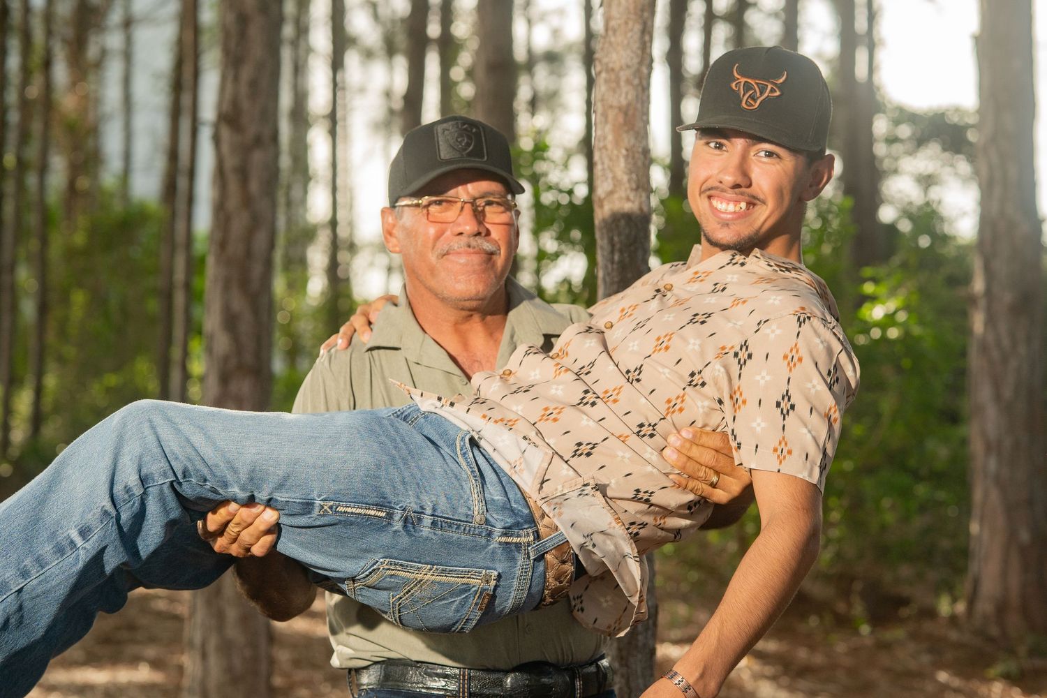 A young Marine in uniform is lifted by an older man in a forest setting, both smiling joyfully.