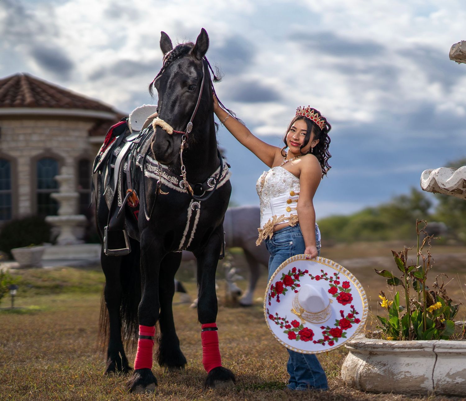 A black horse in decorative tack stands beside someone in a festive dress and red floral hat near a garden.