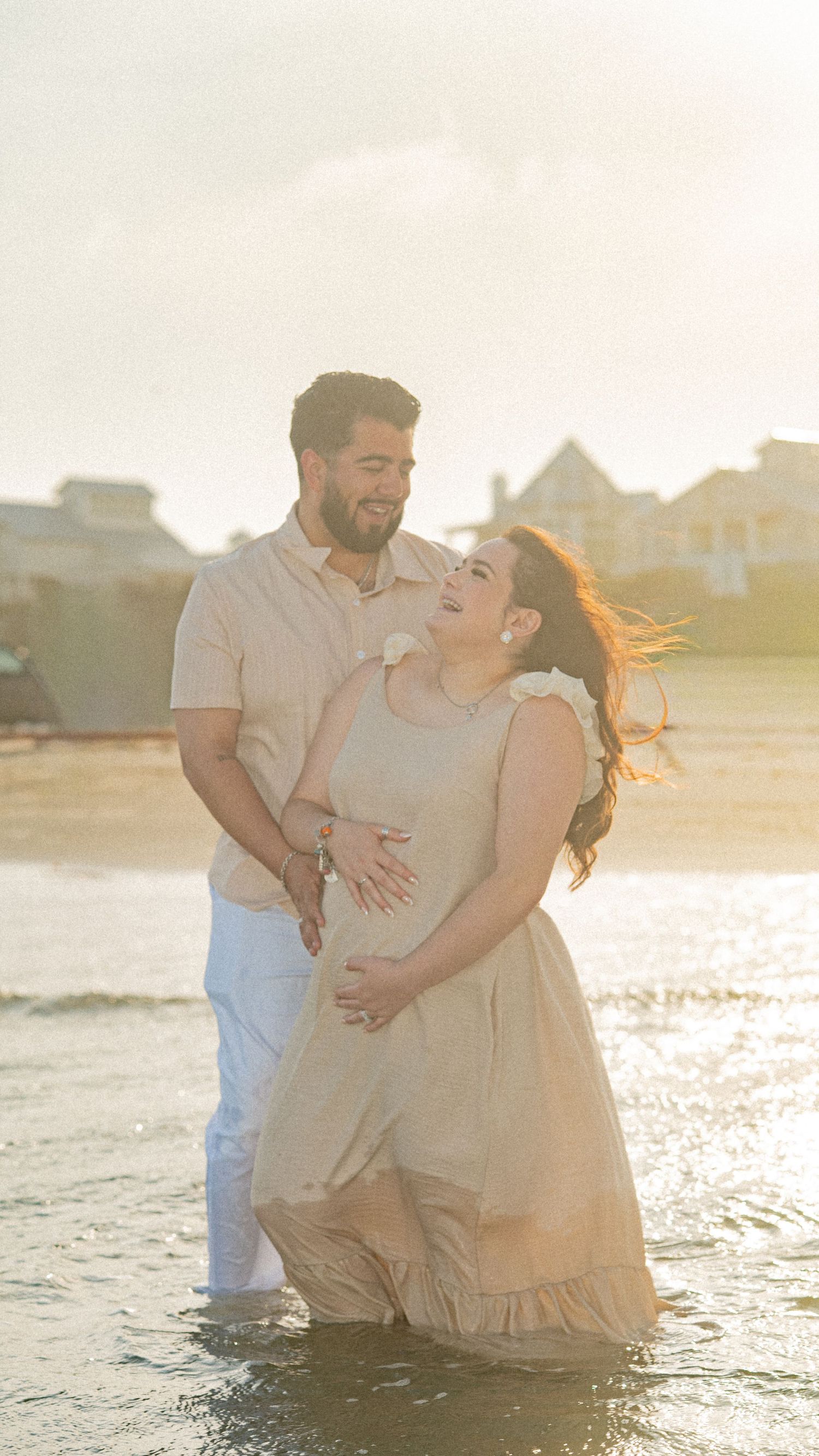 Couple embracing on beach at golden hour, woman in flowing dress cradling baby bump, waves at their feet.