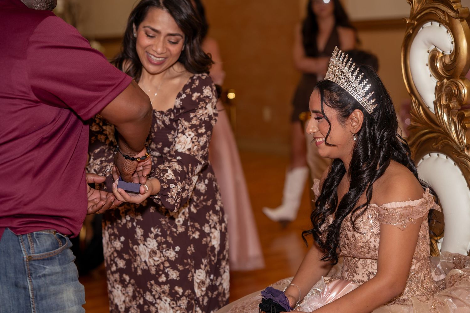 Young woman in tiara and pink quinceañera gown seated while receiving a gift from family members.