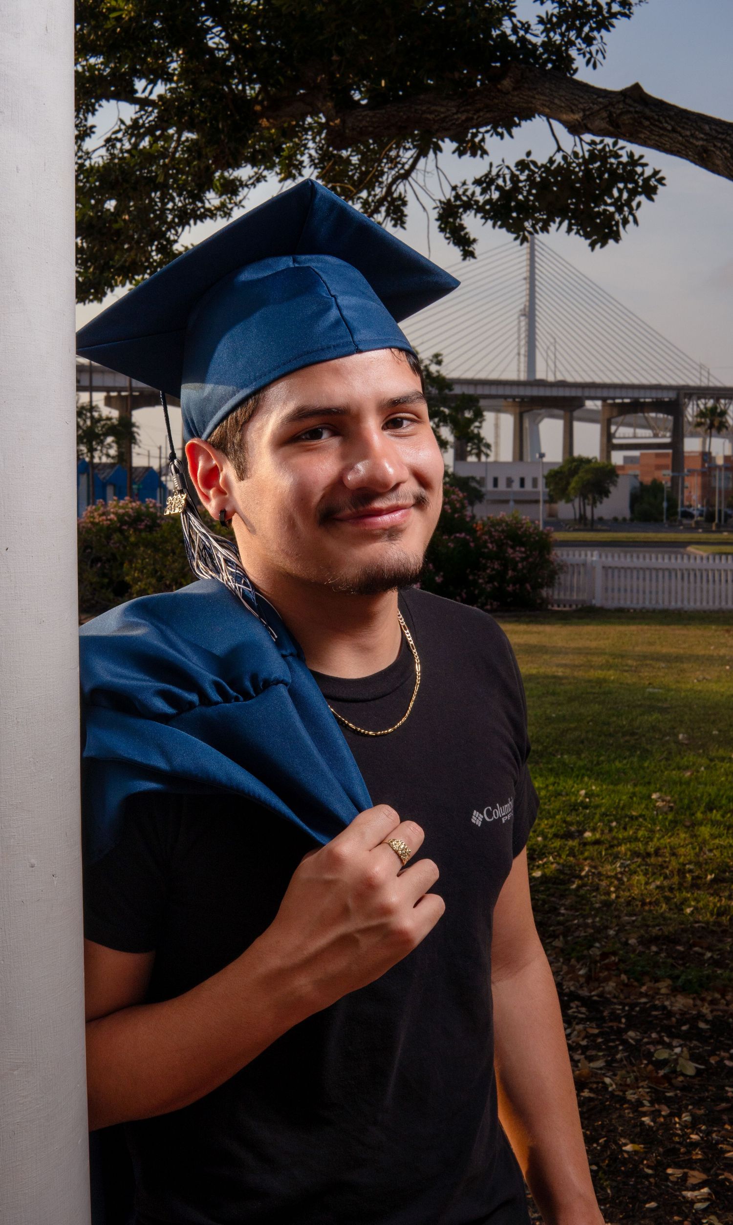 A graduate in blue cap and gown stands outdoors near a bridge with Columbia Sportswear attire.