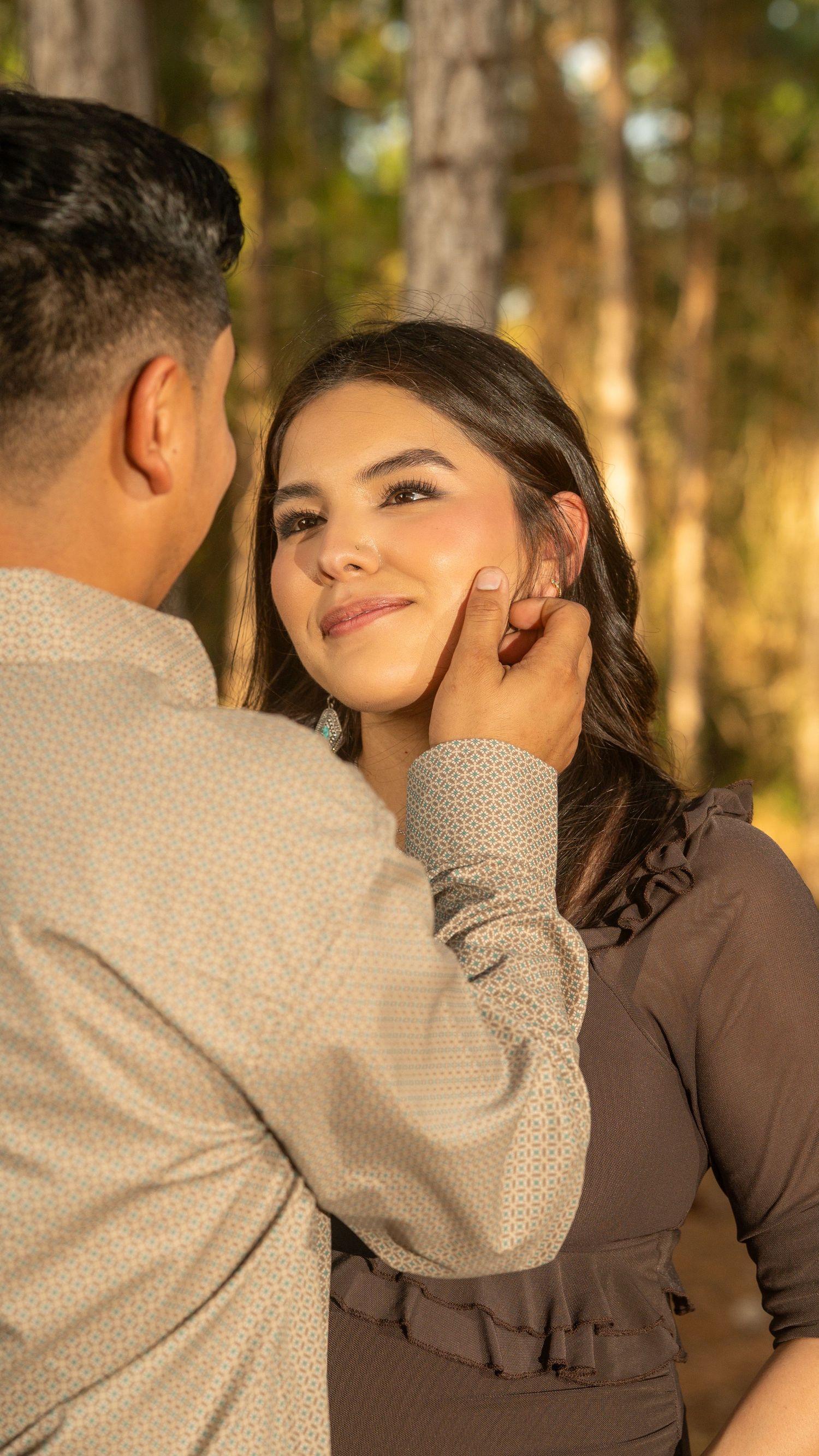 A man gently cups a smiling woman's face in a romantic outdoor setting among trees in warm golden light.