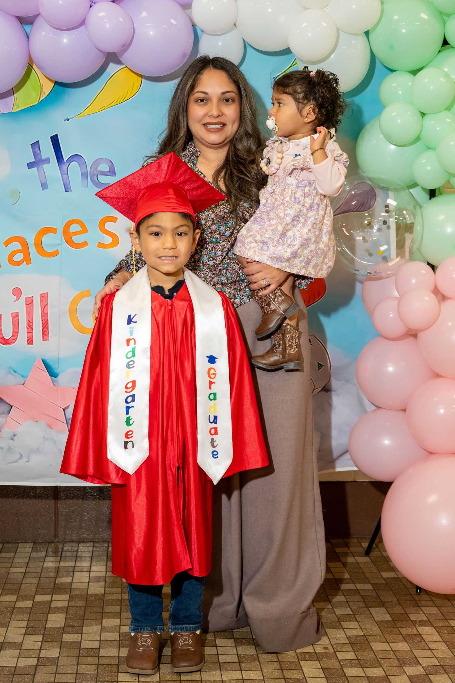 A proud mother holds her toddler beside her kindergarten graduate in red cap and gown at ceremony.