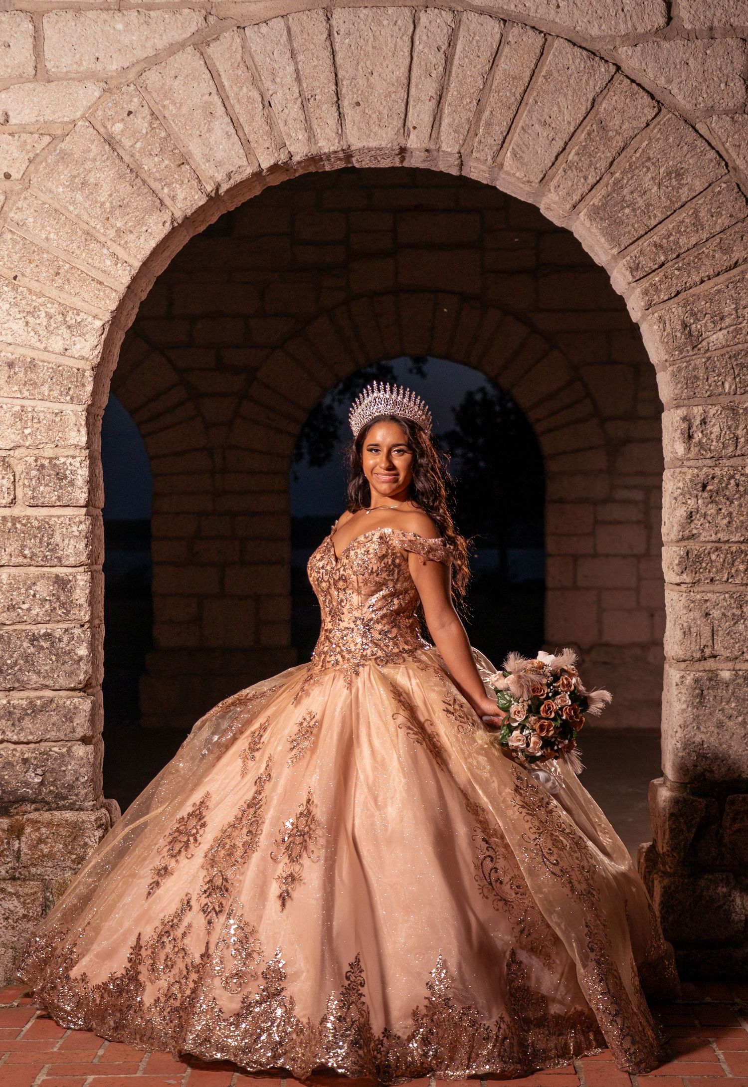 A stunning champagne-colored ball gown with detailed embroidery photographed under a stone archway.