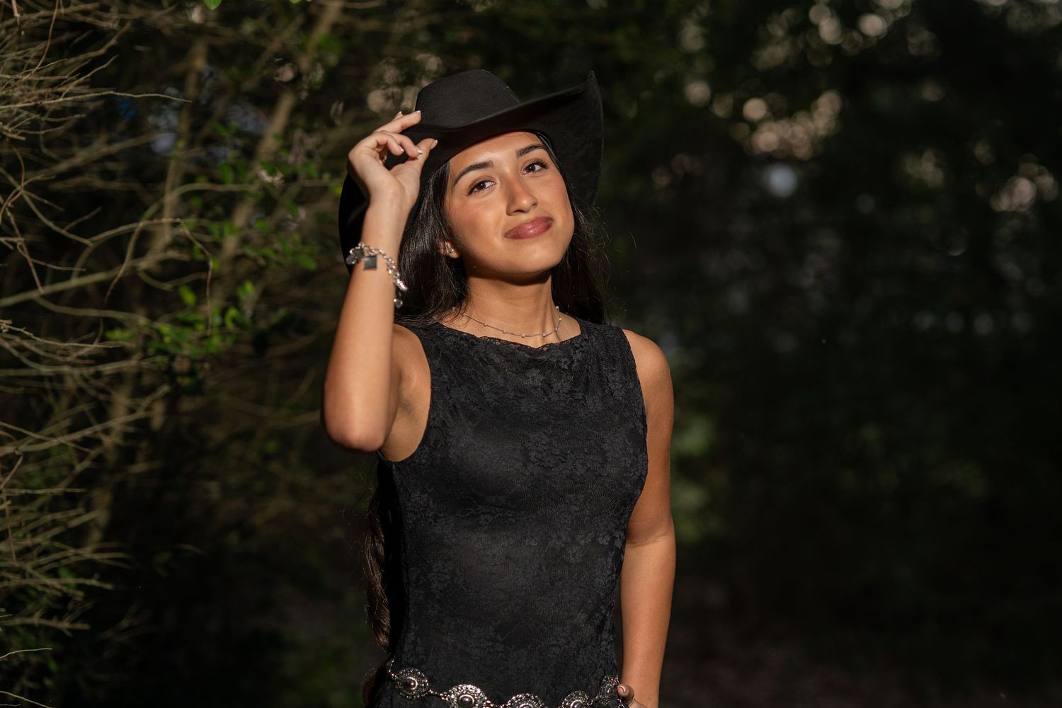 Young woman in black lace dress and black hat posing outdoors among trees in warm golden light.