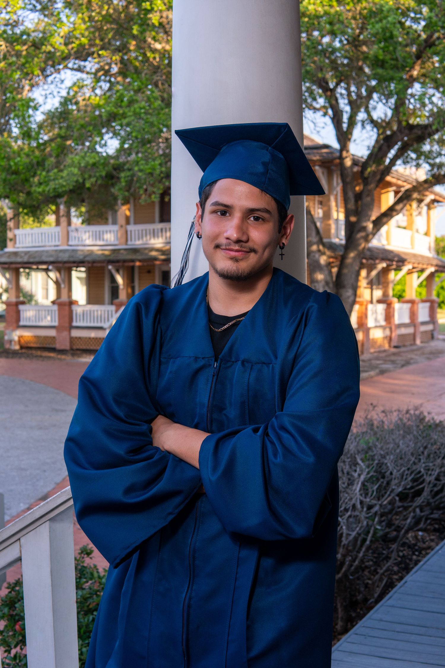 A graduate in navy blue cap and gown stands on a porch with trees and buildings in the background.