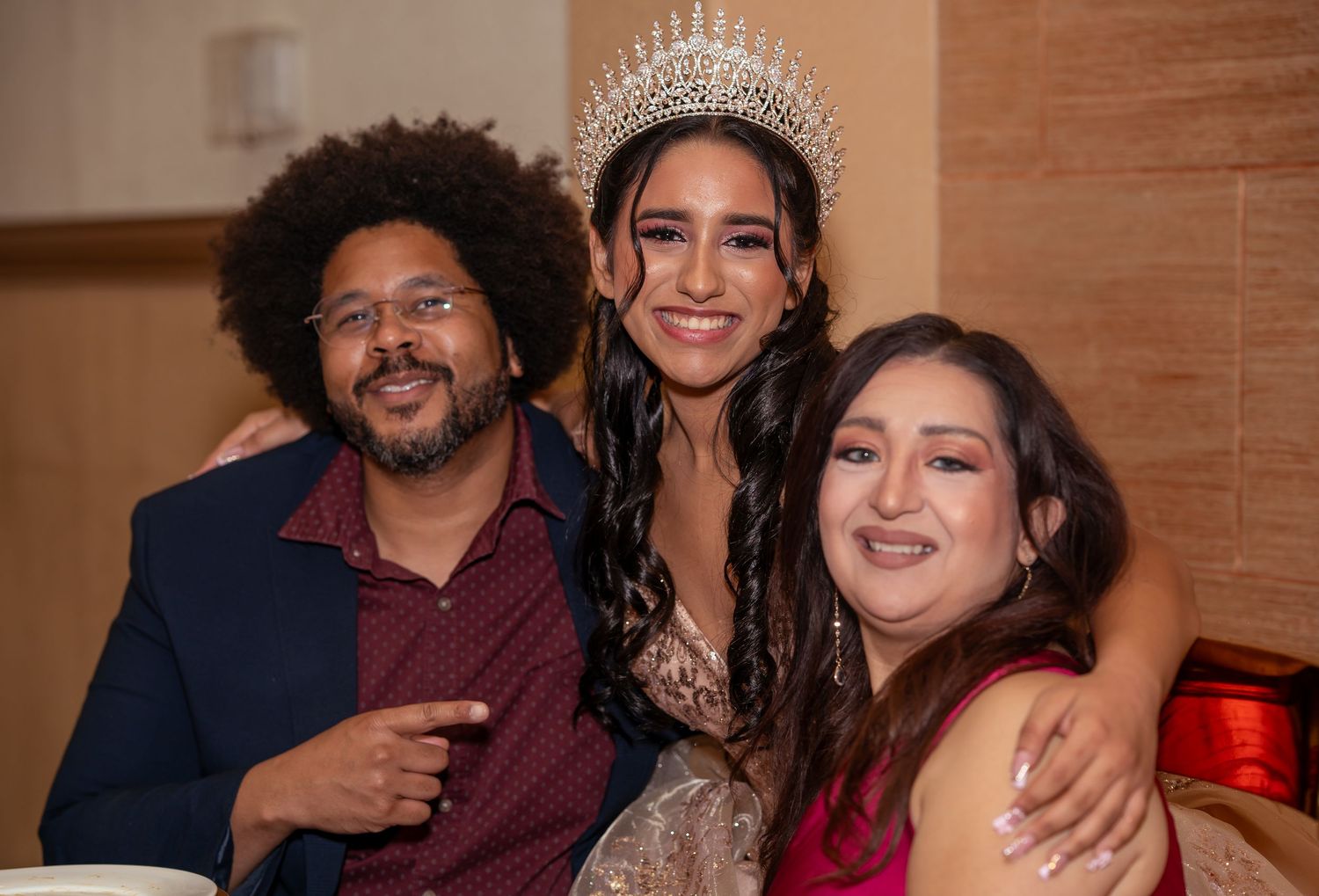 Quinceañera girl in rhinestone crown smiles with two friends at her birthday celebration.