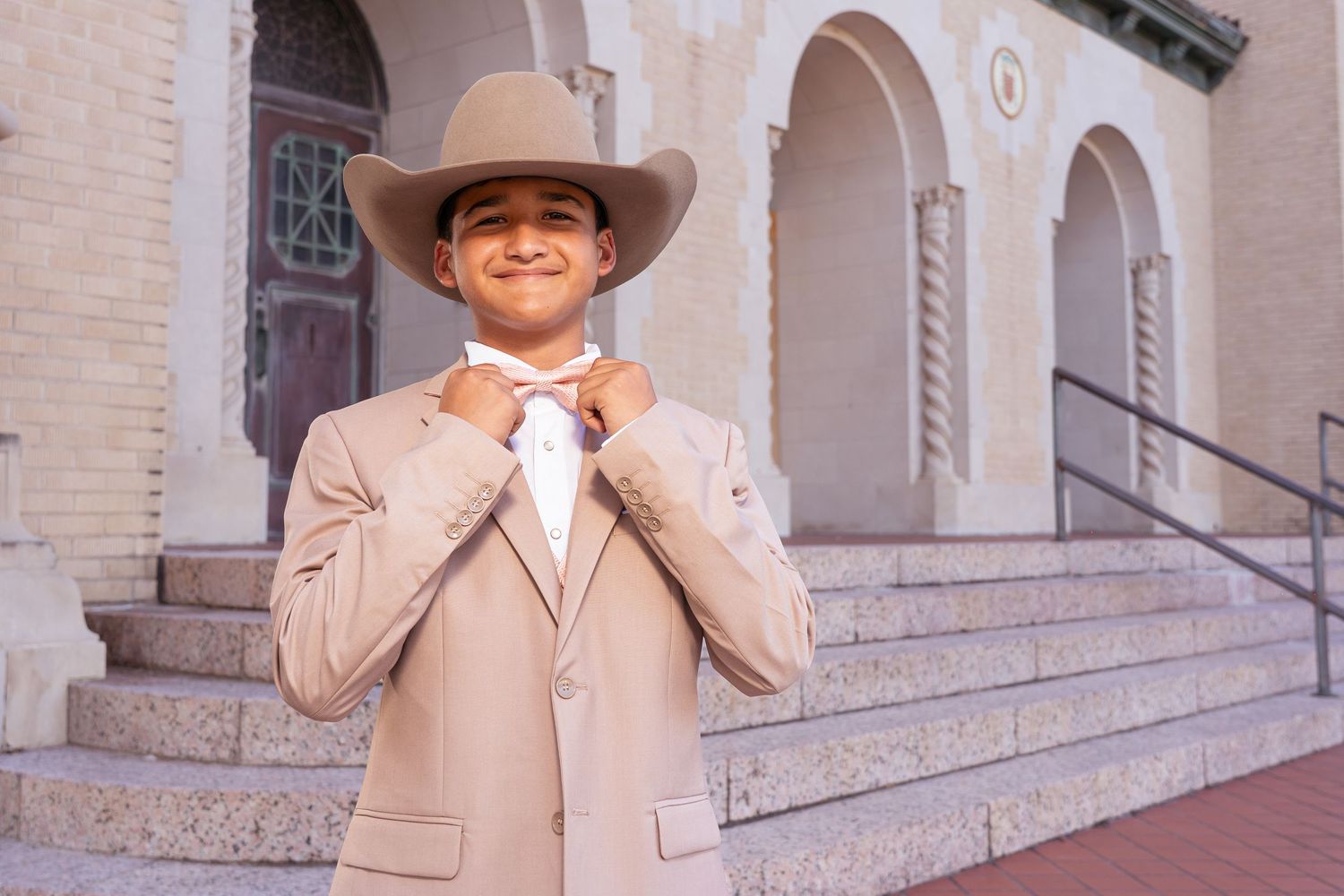 Person in western attire poses confidently on stone steps with arched columns in the background.