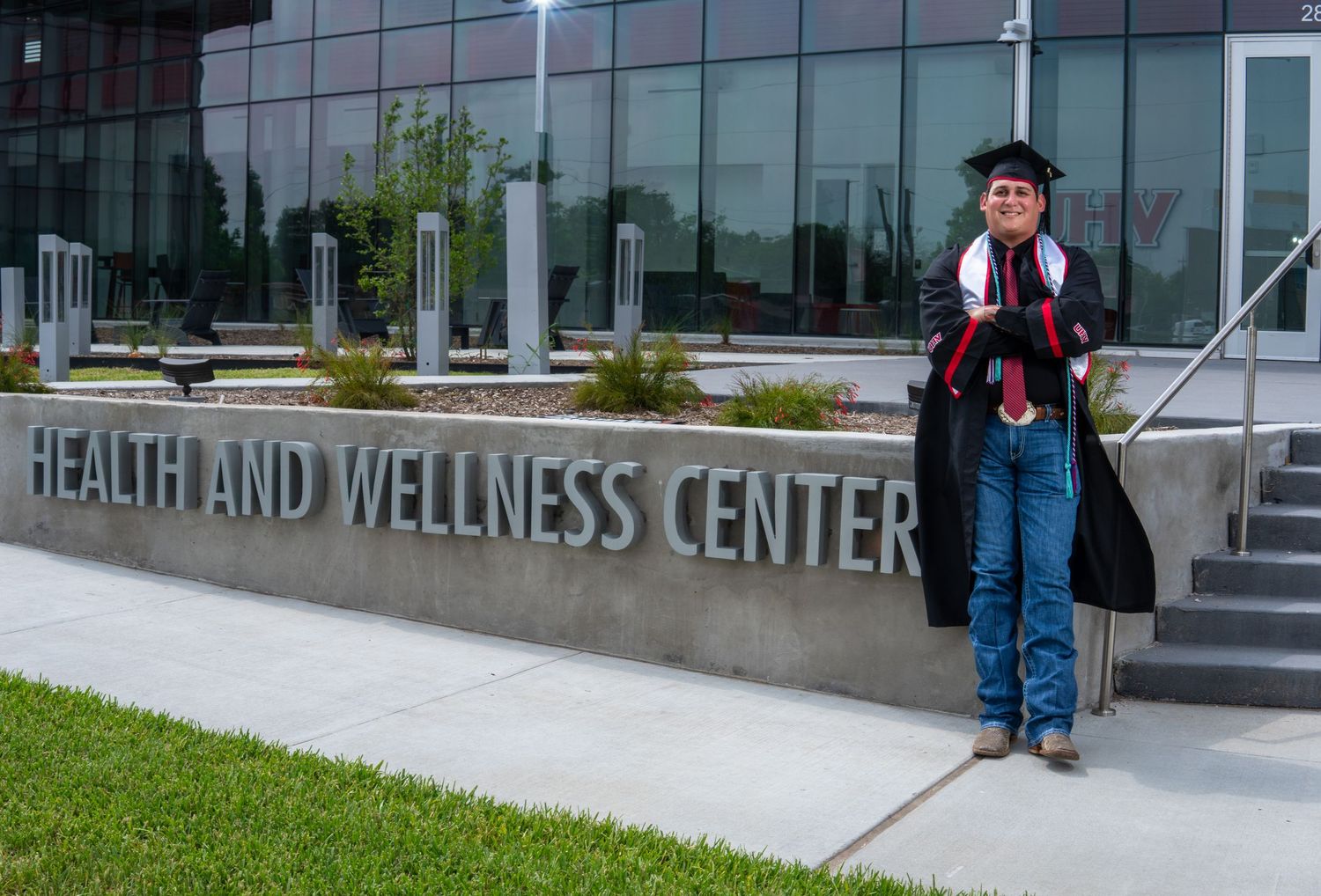 A graduate wearing a cap, gown and backpack stands on the steps of a Health and Wellness Center building.