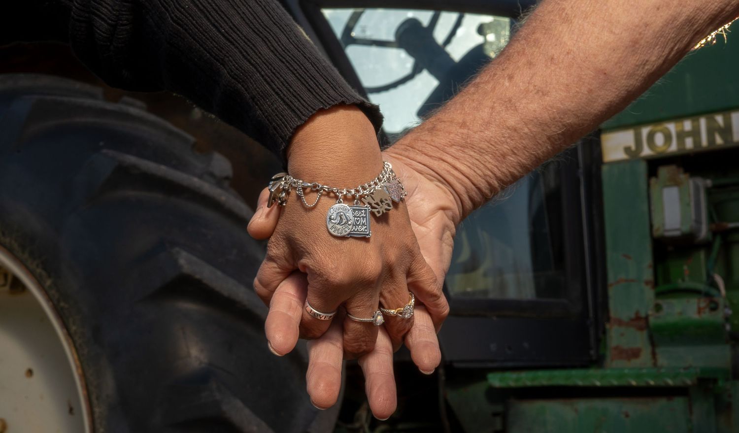 Two hands clasped together, one wearing a silver charm bracelet and rings, near a green John Deere tractor tire.