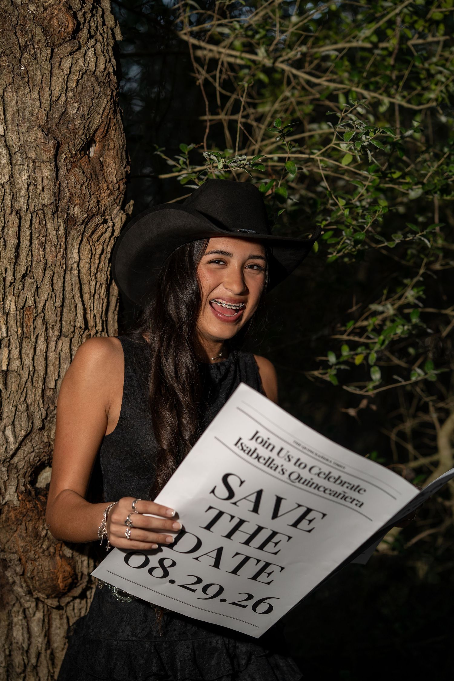 Teen girl in black dress and cowboy hat holds quinceañera Save the Date sign by a tree.