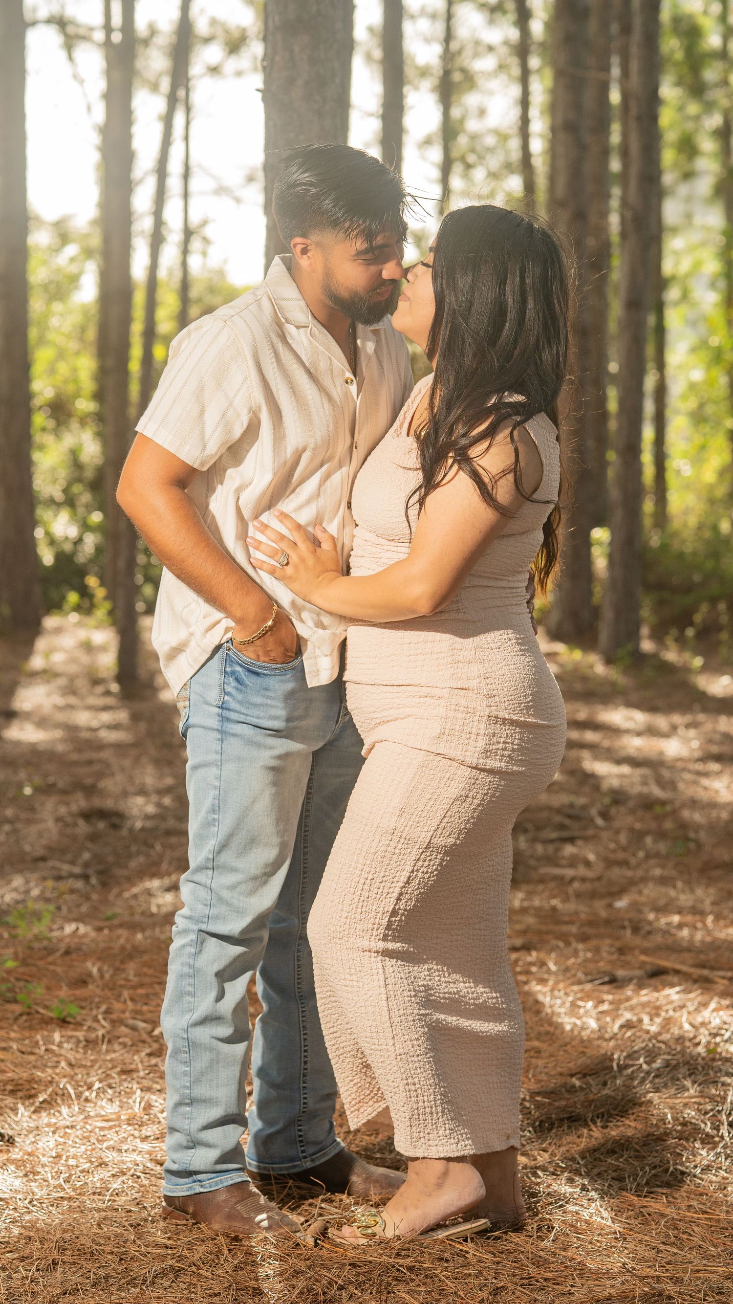 Couple sharing a romantic moment in a sunlit pine forest, woman in beige dress and man in jeans and white shirt.