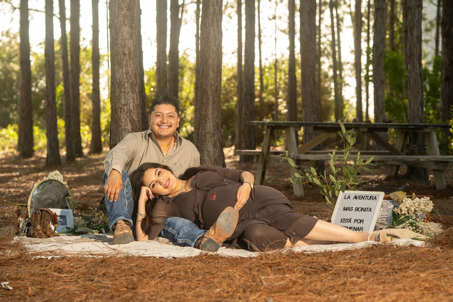 Couple poses in pine forest for maternity photoshoot, woman resting on mans lap on blanket among trees.