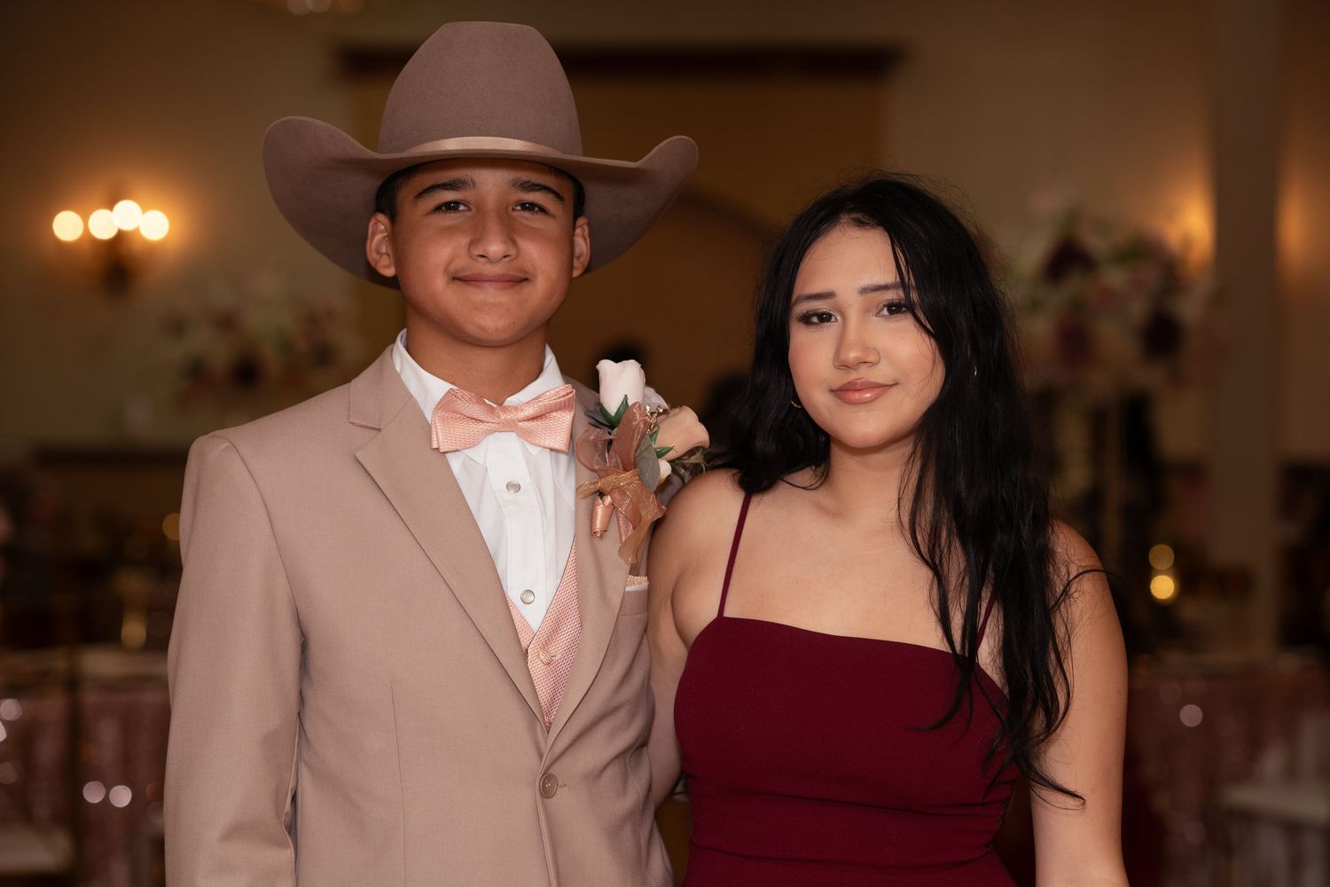 Teen boy in tan suit and cowboy hat posing with girl in burgundy dress at a quinceañera celebration.