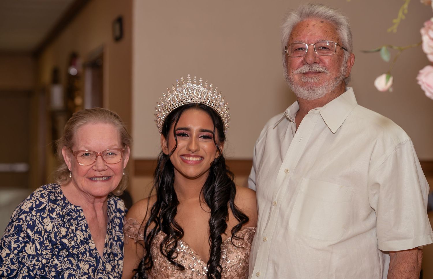 Young woman wearing a rhinestone crown poses with elderly grandparents at a quinceañera party.
