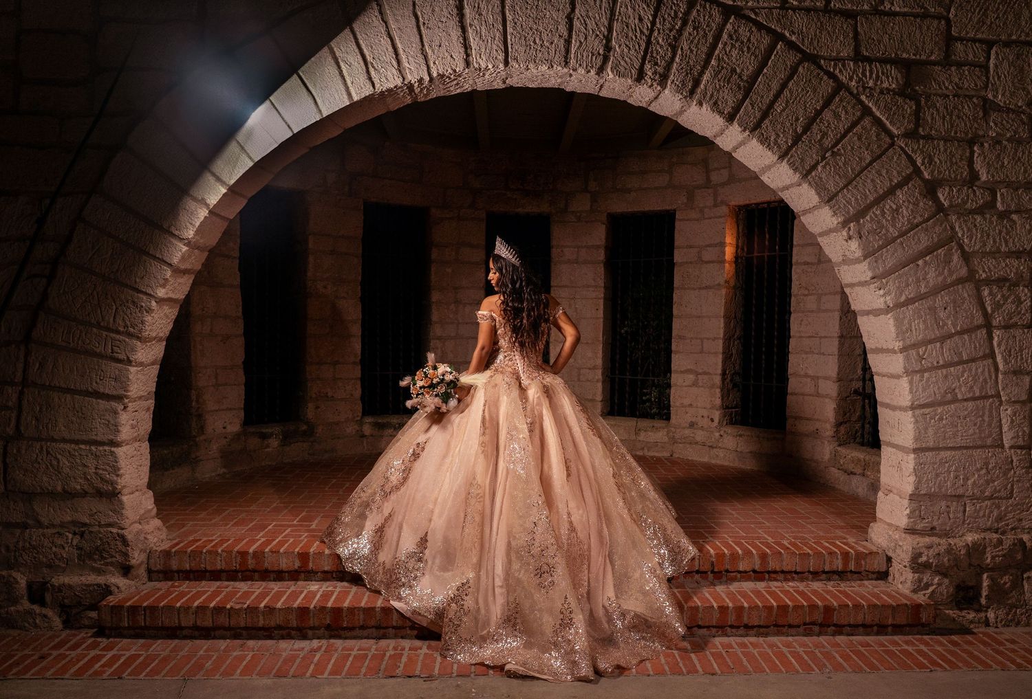 A stunning bride in a flowing champagne ballgown poses beneath a dramatic brick archway at night.