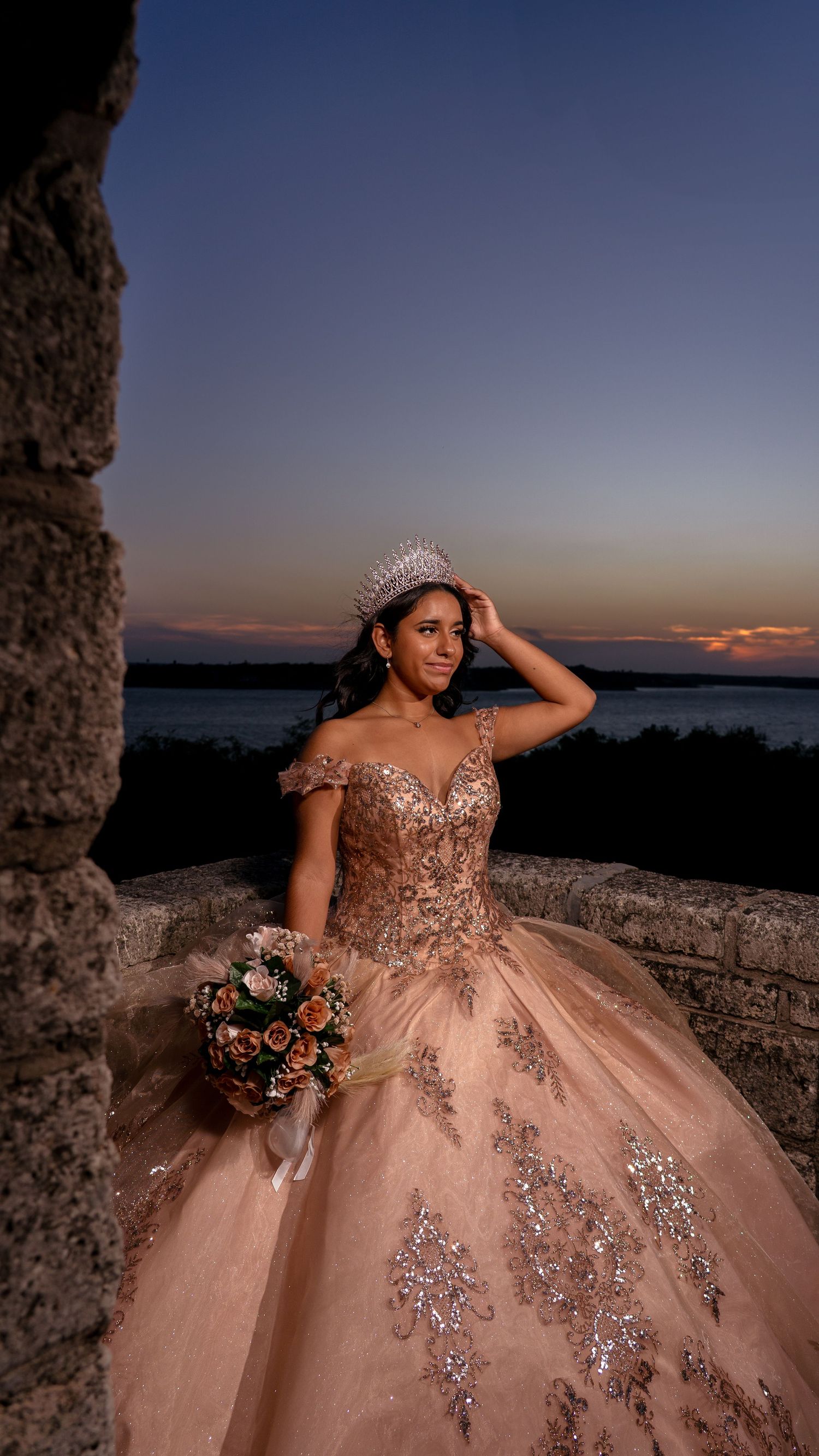 Elegant quinceanera dress in blush pink with sparkling details and tiara photographed at sunset against stone architecture.