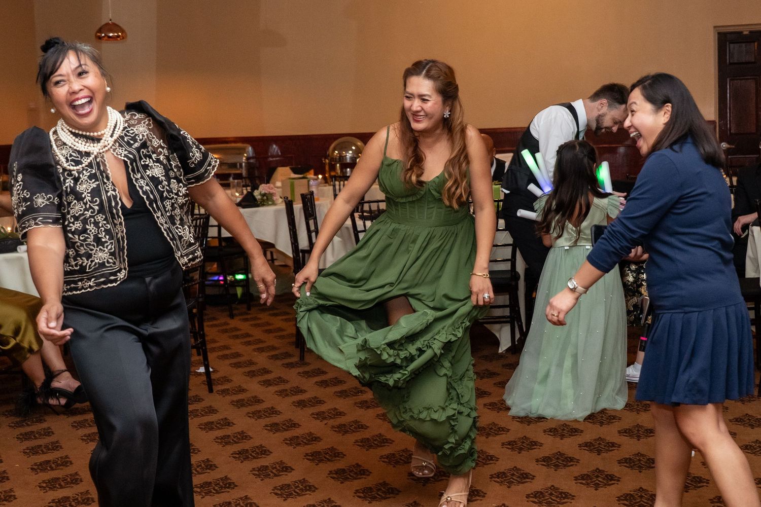 Three women laugh and dance together in a ballroom during a festive wedding reception celebration.