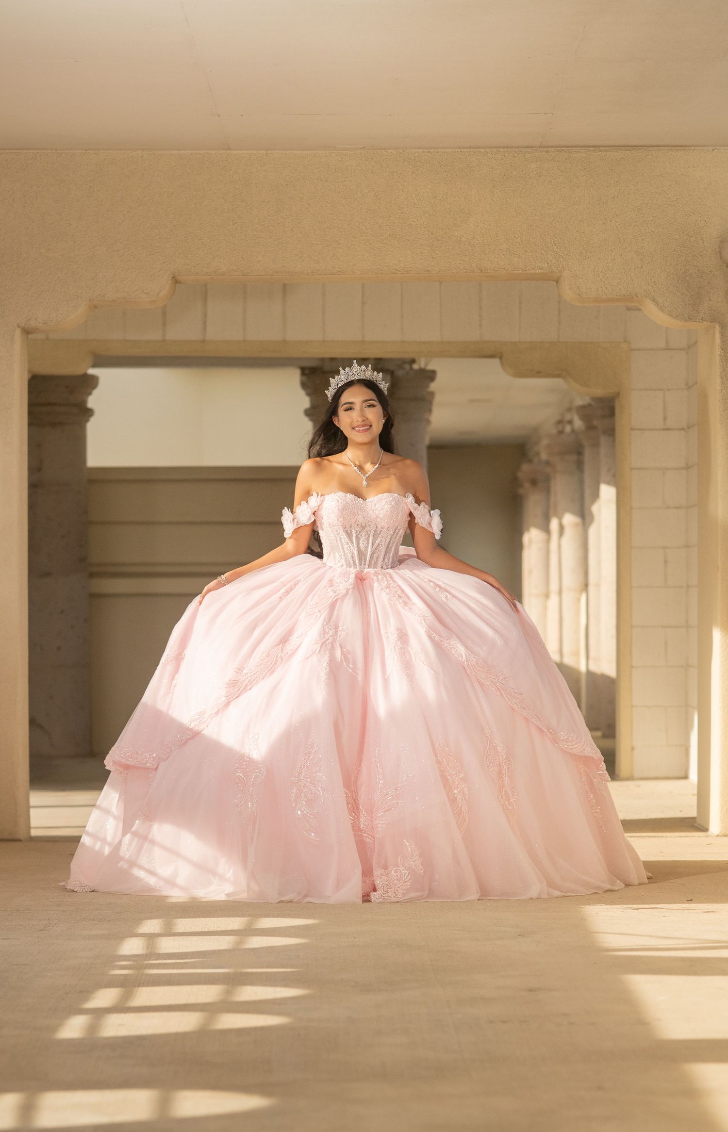 Young woman in a pink ball gown and tiara poses elegantly for a quinceañera photo in a sunlit archway.