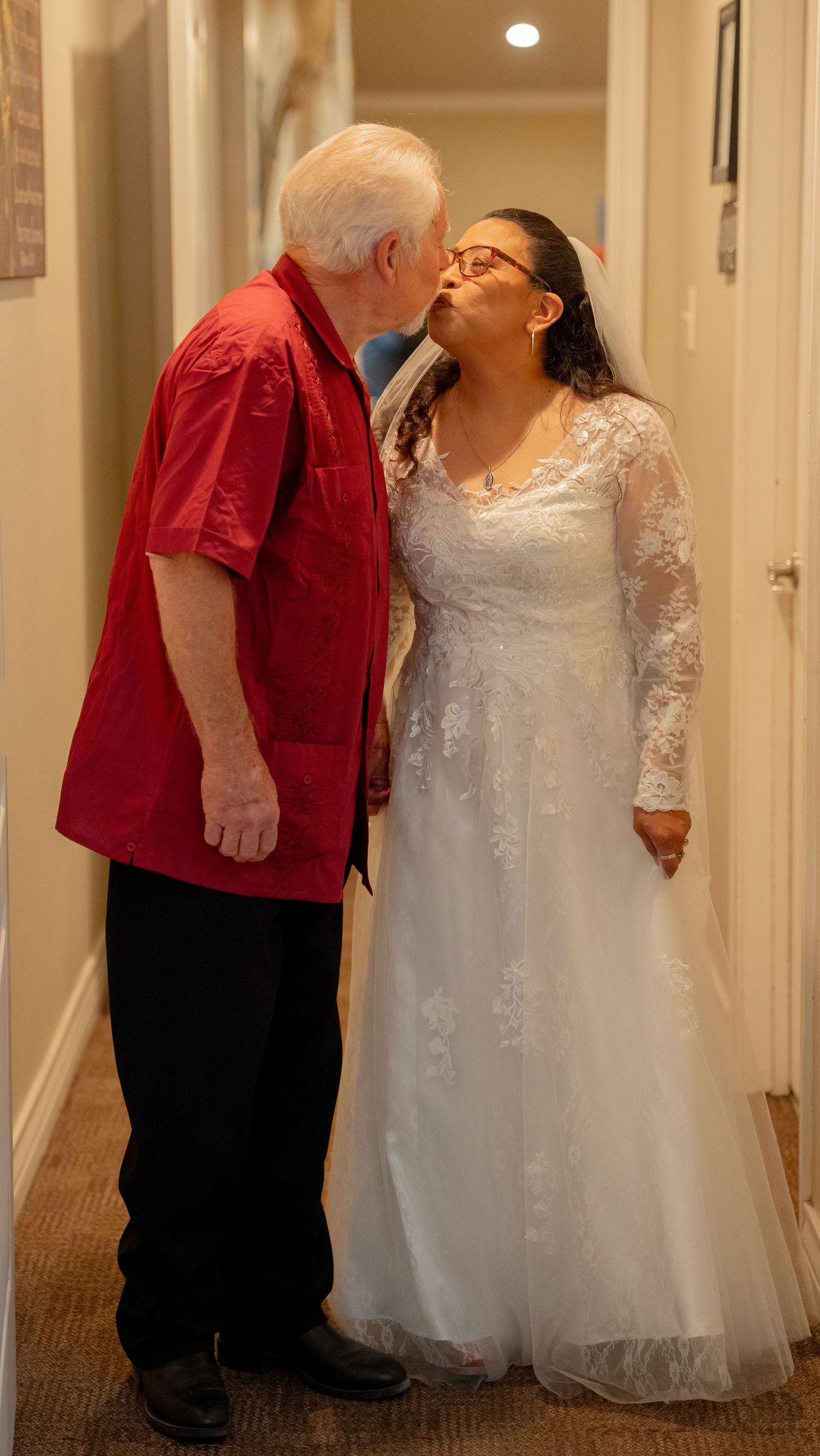 Elderly couple sharing a kiss in a hallway, bride in white lace gown and groom in red shirt on wedding day.