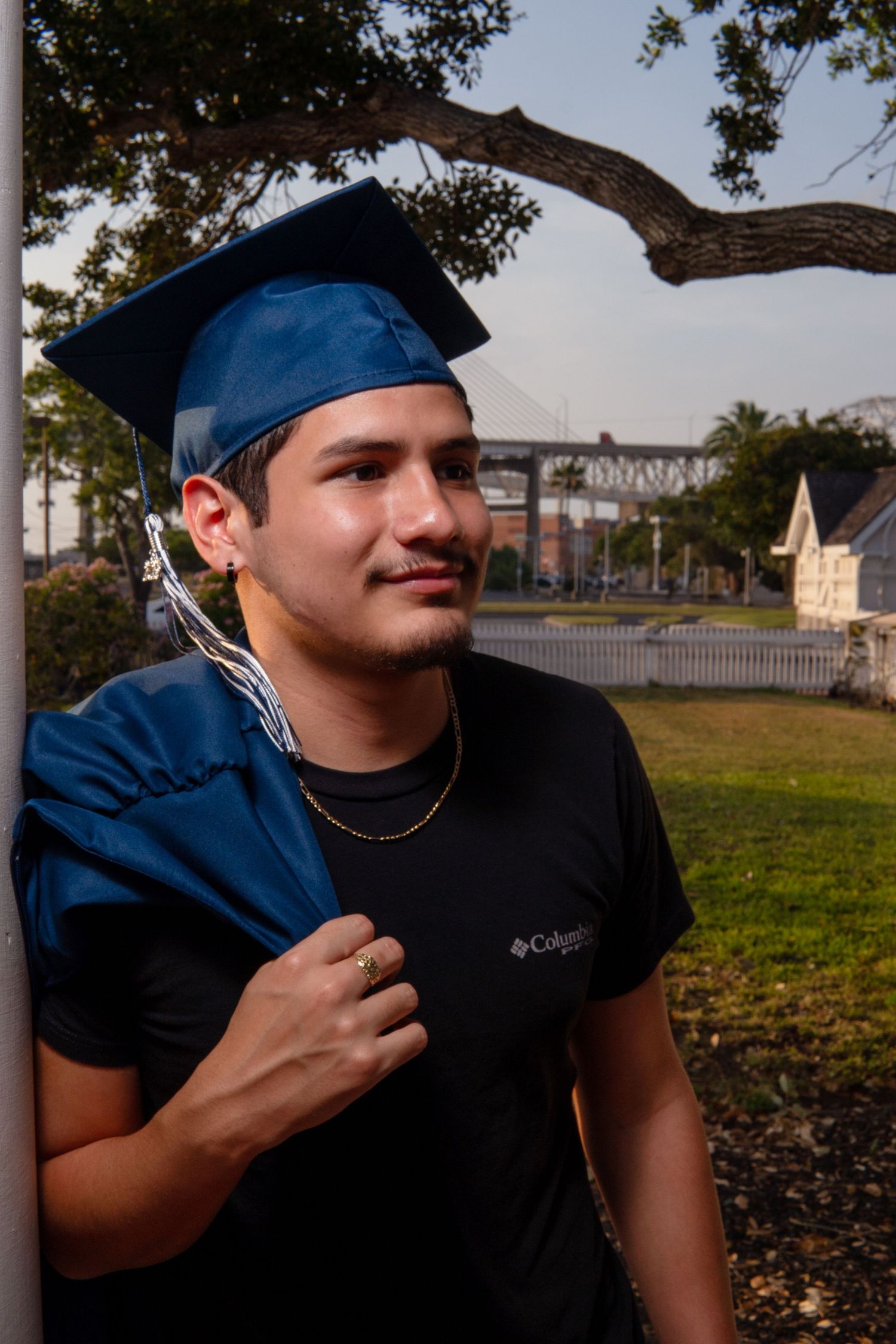Graduate wearing blue cap and black shirt smiles while standing outdoors near a tree at sunset.