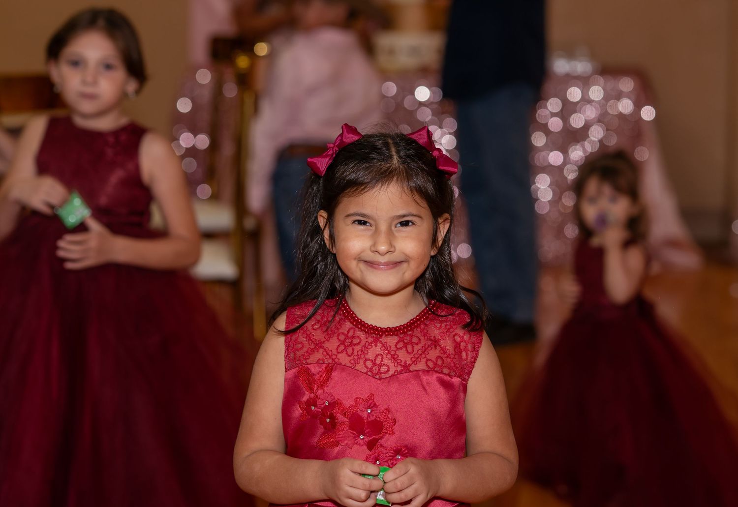 Young girl in red dress smiles at formal event with other children in matching burgundy dresses in background.