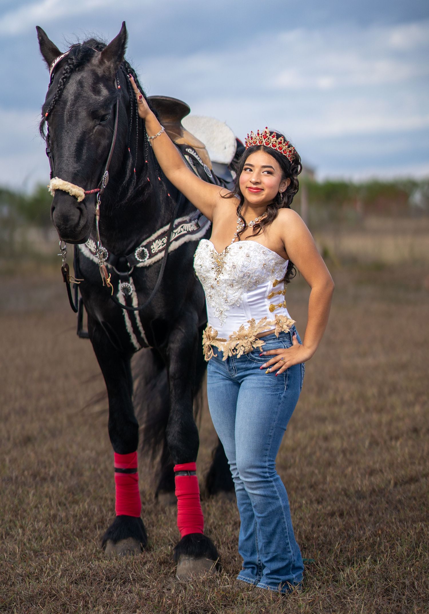 A black horse wearing red leg wraps stands in a field with someone in jeans and a white lace top.