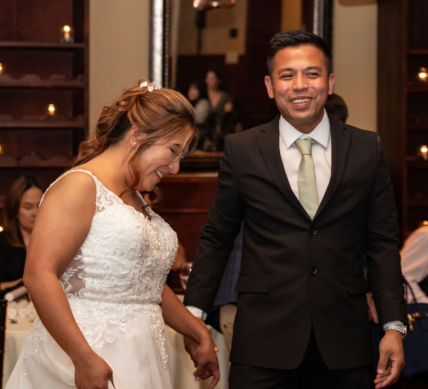 Joyful bride in white lace gown and groom in dark suit with green tie laughing together at wedding reception.