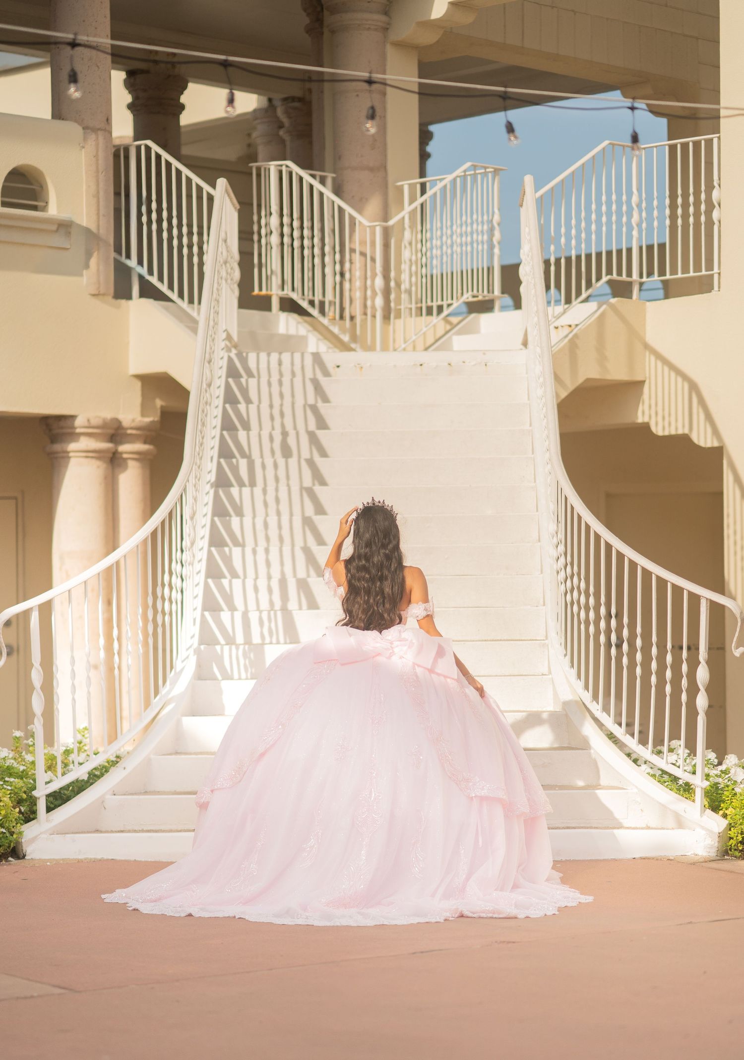 Young woman in a pink ball gown quinceañera dress poses on white grand staircase from behind.