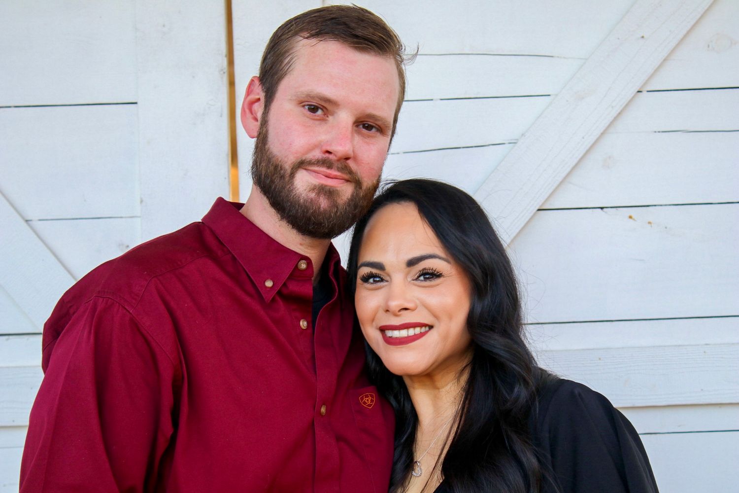 A couple poses in front of a white wall wearing burgundy and black.