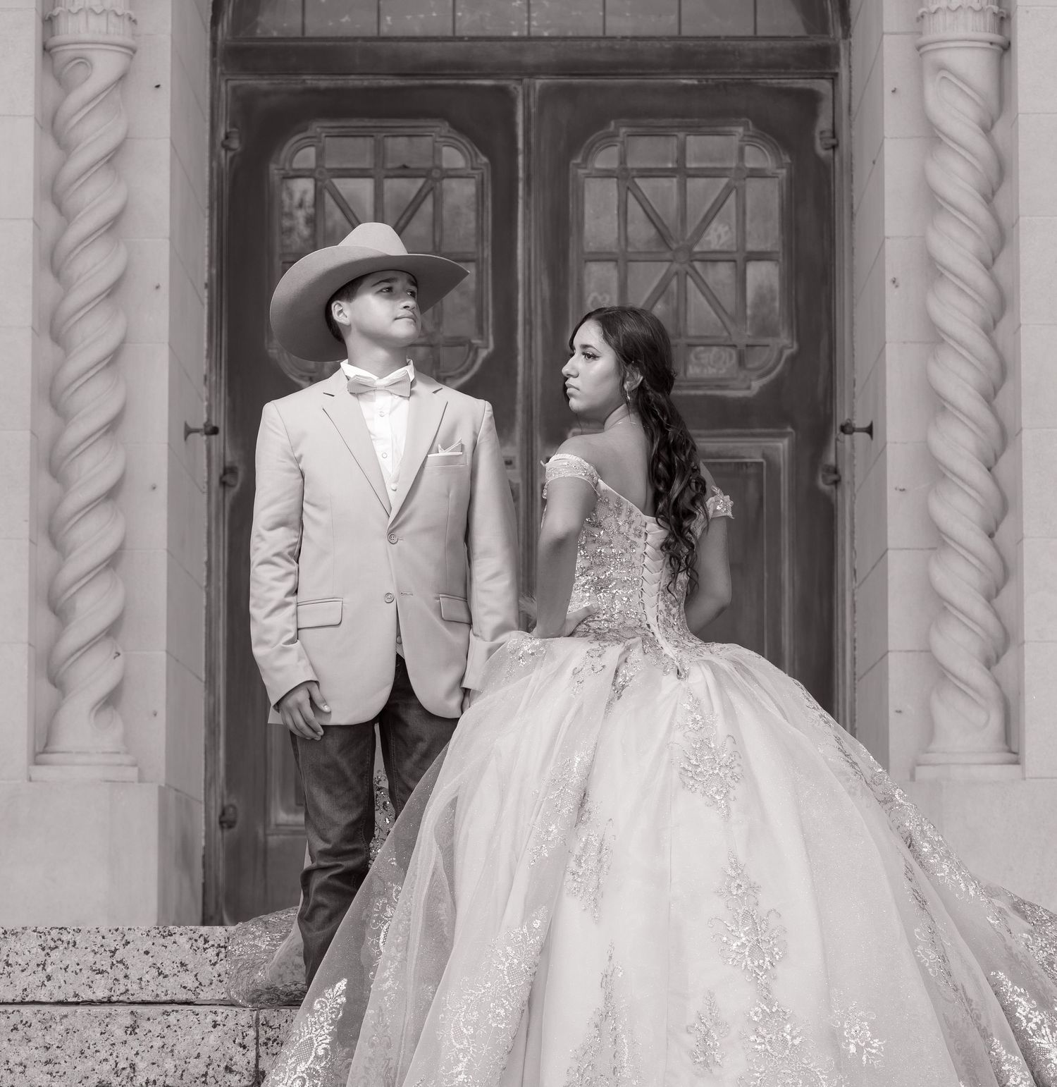 A couple poses in formal attire for a quinceañera photo shoot in black and white against an ornate doorway.