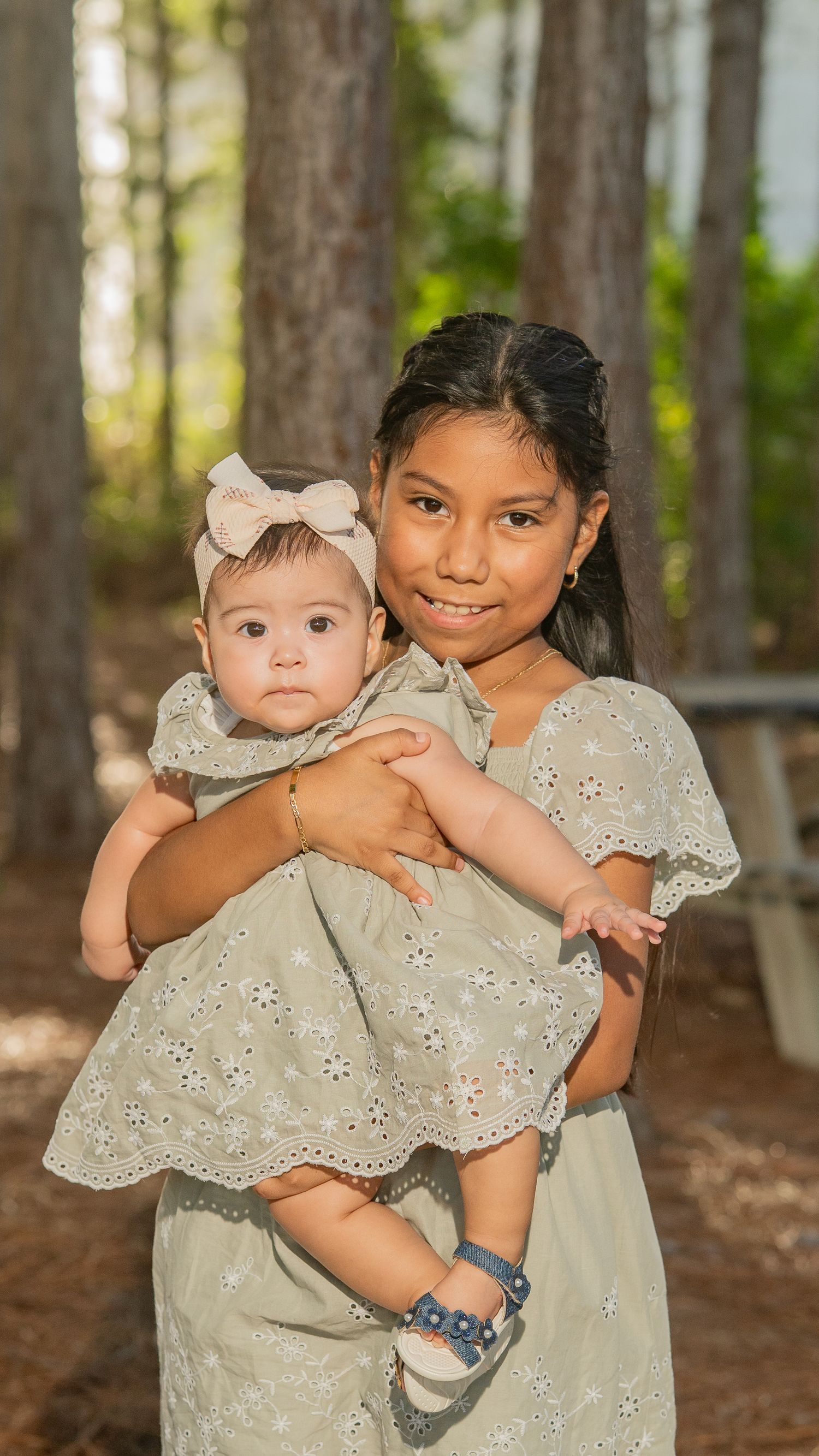 Older girl in sage dress holds baby sister with bow headband in a forest setting.