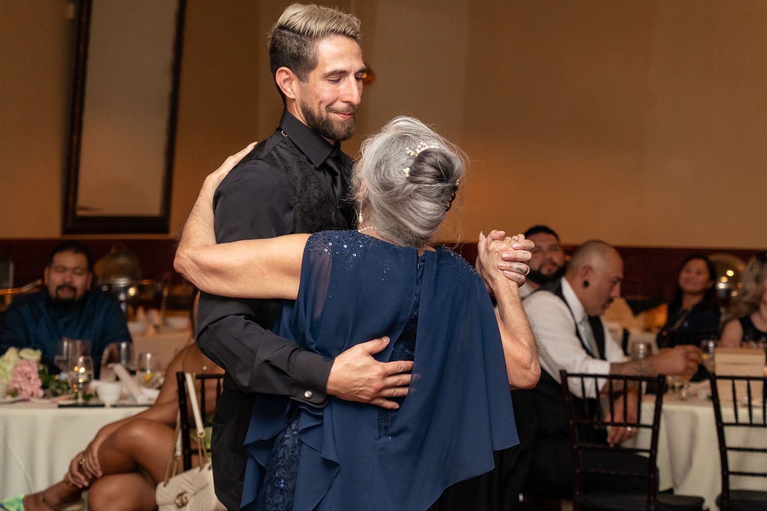 A man in a black shirt shares a dance with an older woman in blue at a formal reception.