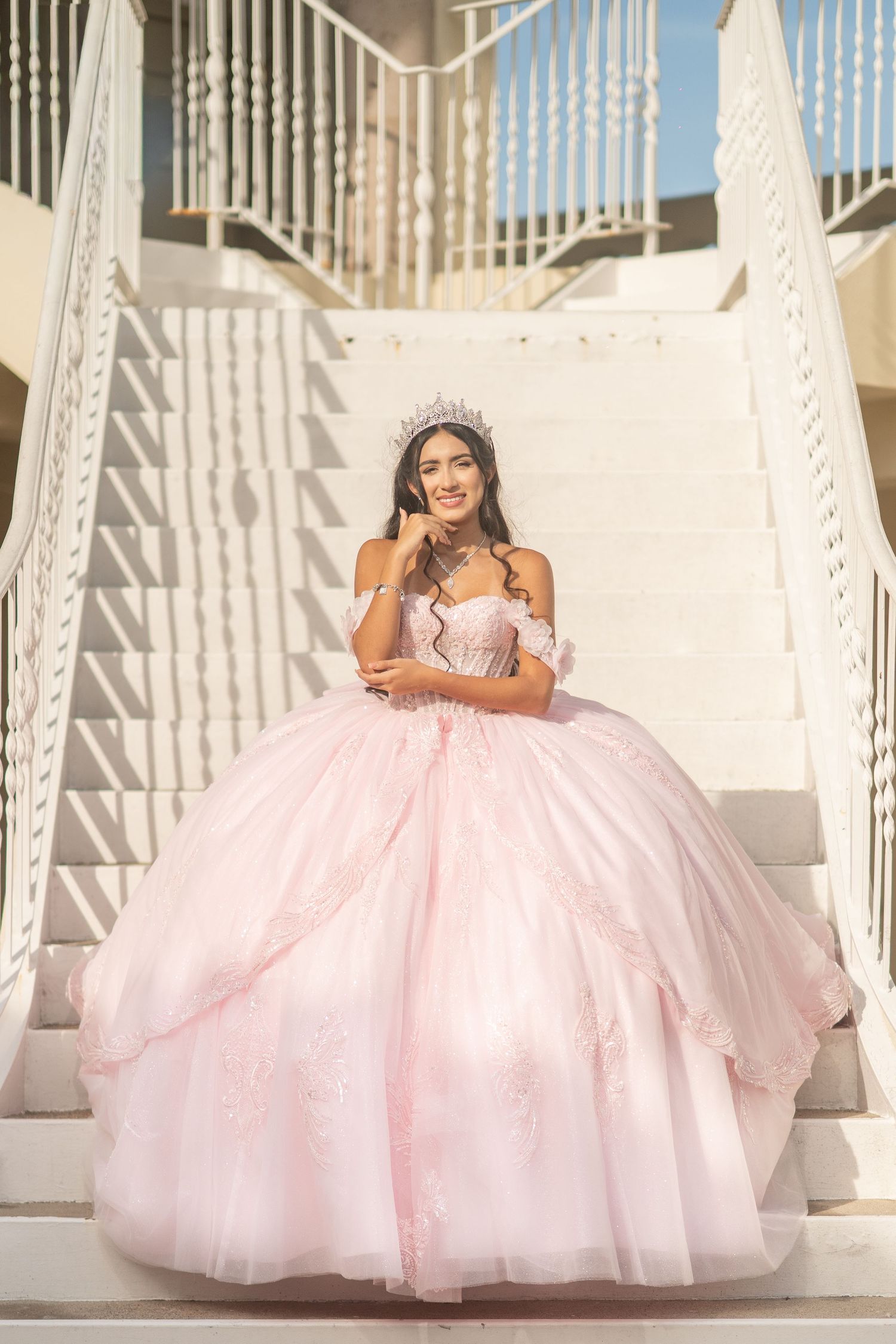 Young woman in a pink ball gown and tiara poses gracefully on white stairs during a quinceañera celebration.