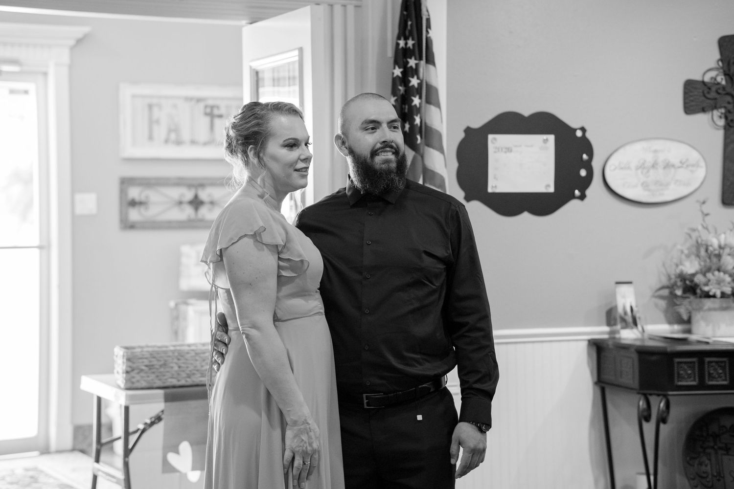 Black and white photo of couple standing together indoors near American flag at casual wedding.