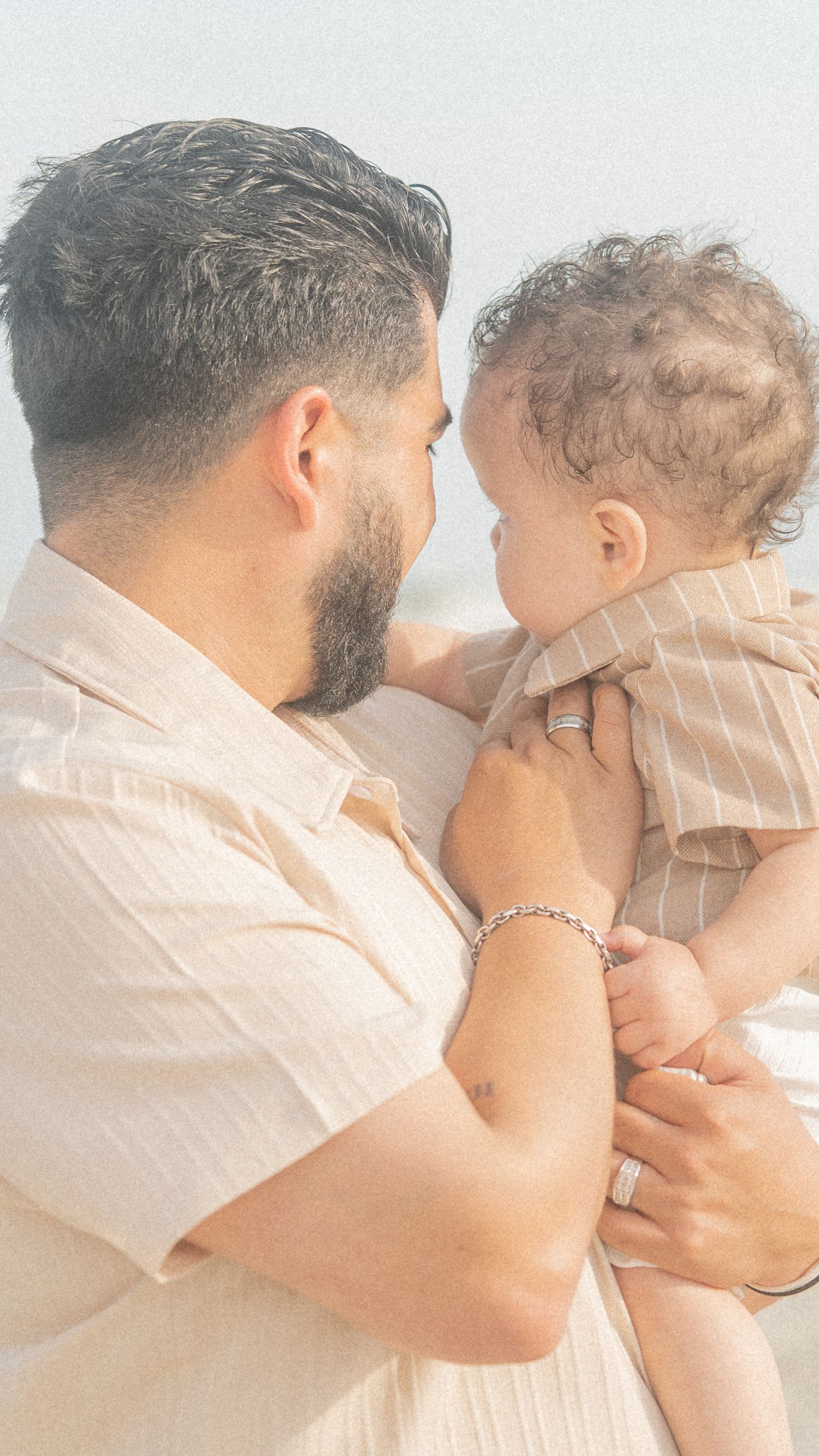 A bearded father in a beige shirt tenderly holds his curly-haired baby, sharing a gentle nose-to-nose moment.