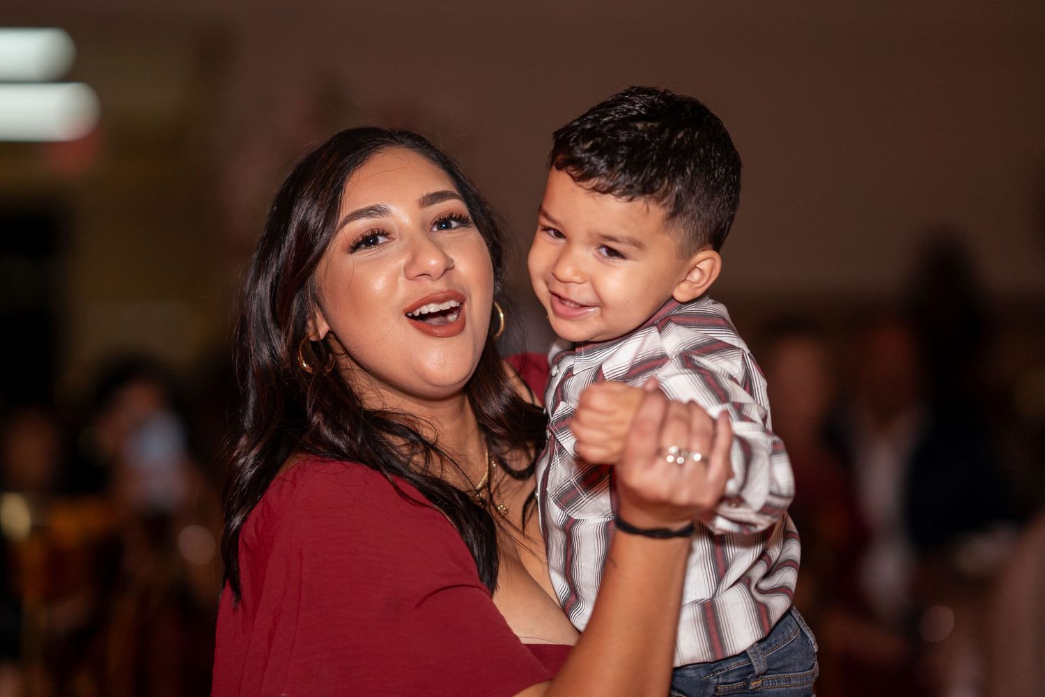 Joyful woman in red top holds a smiling toddler boy at a festive celebration event.