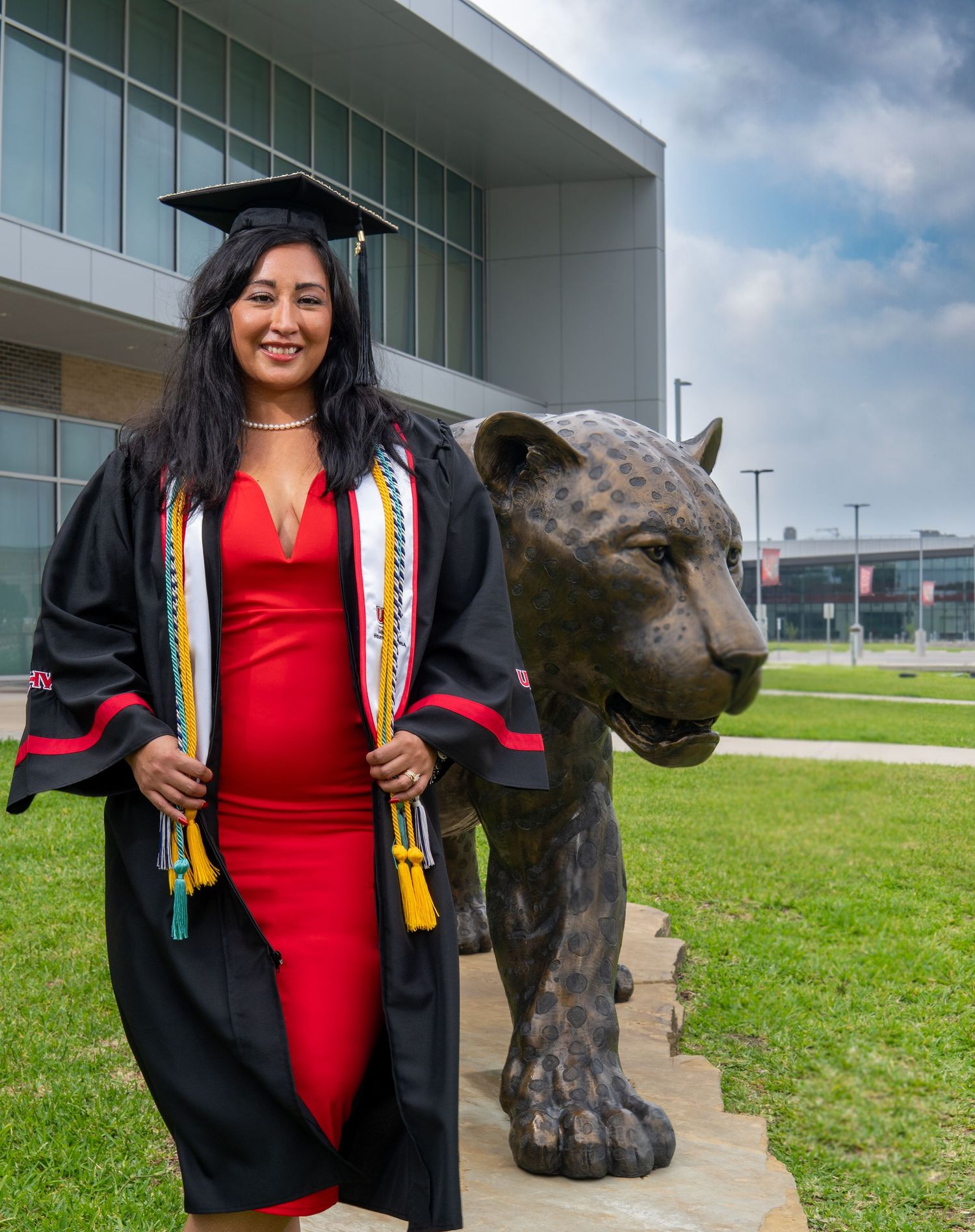 Graduate in cap and gown poses next to large bronze panther statue on university campus lawn with modern building behind.