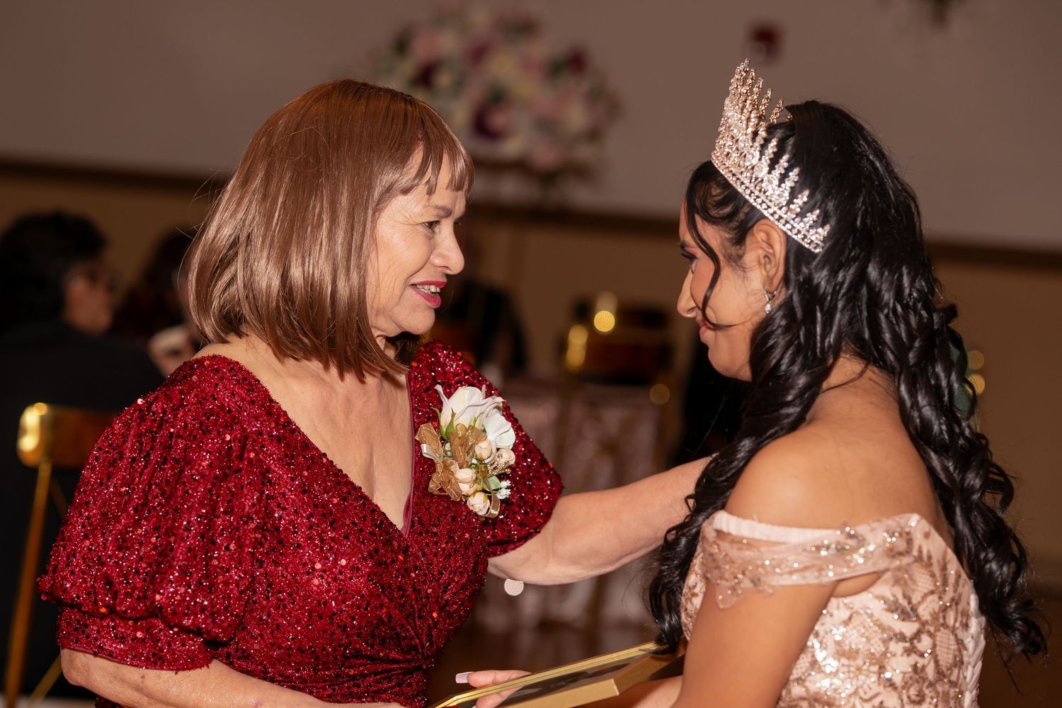 A quinceañera girl in a rose gold gown and tiara shares a tender moment with an older woman in red.