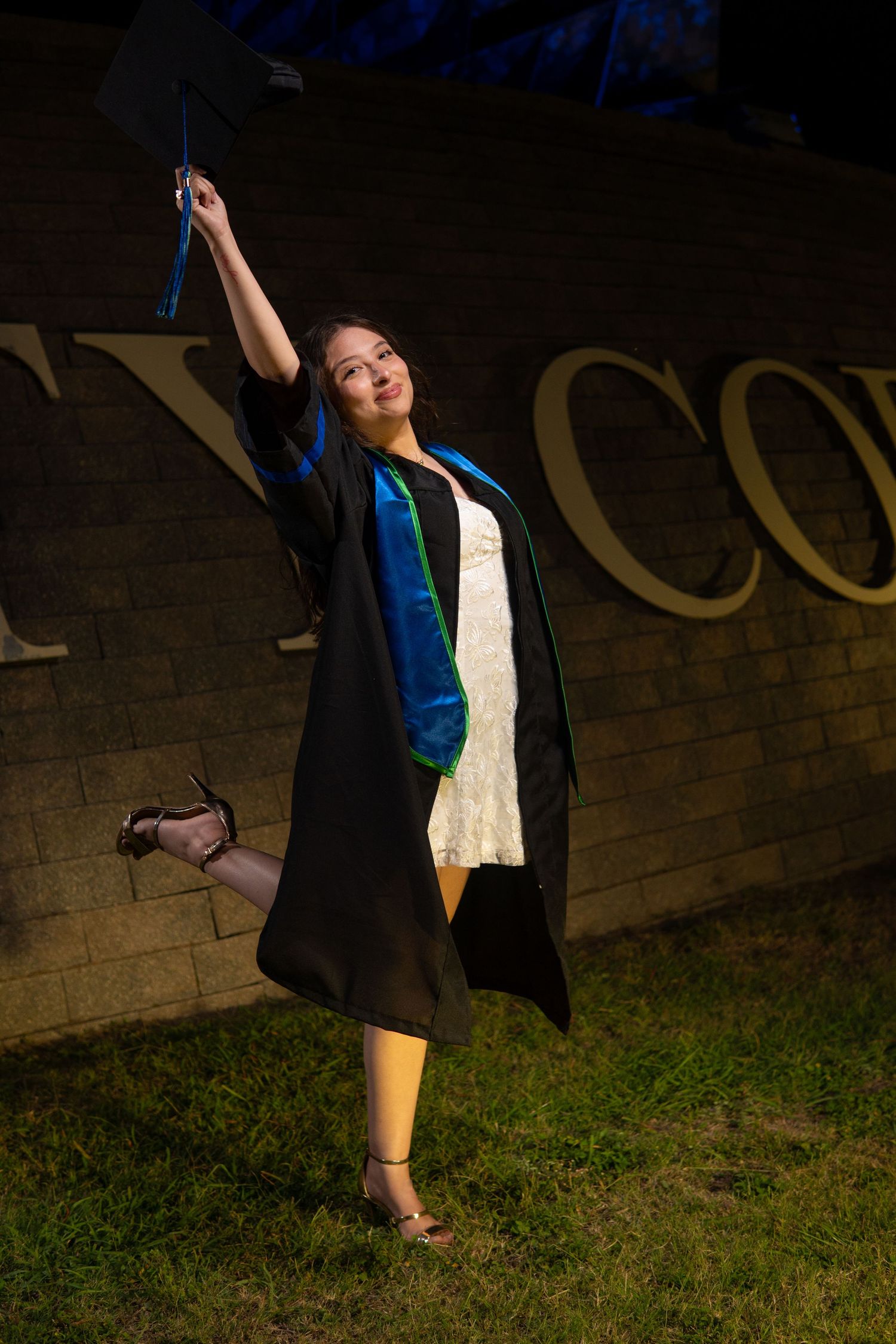 Graduate in black cap and gown with blue stole strikes a joyful pose against a brick wall on grass.